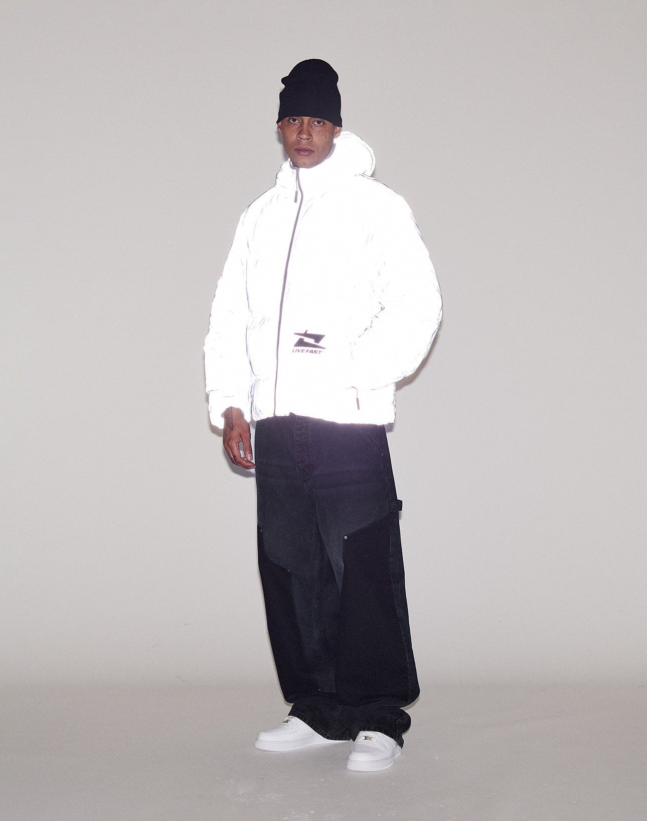 A person wearing a LIVE FAST DIE YOUNG Live Fast Puffer Jacket, black beanie, loose black pants, and white sneakers stands against a plain light-colored background.