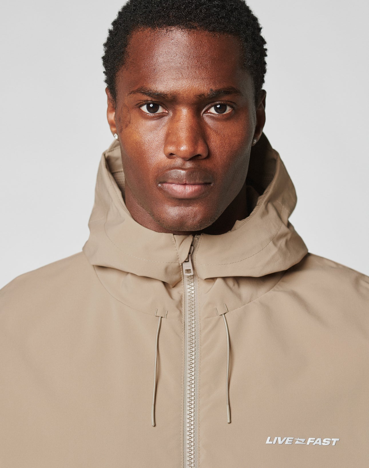 A man with short curly hair wears the LIVE FAST DIE YOUNG Basic Shell Jacket in beige, featuring an oversized fit, water-repellent zipper, and LIVE FAST in reflective print on the chest, looking at the camera with a neutral expression.
