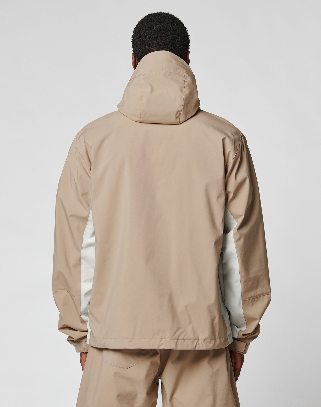 A person wears the LIVE FAST DIE YOUNG Basic Shell Jacket in beige with weiß panels and oversize fit, standing facing away against a plain light background. The jacket includes a wasserabweisenden Reißverschluss for added functionality.