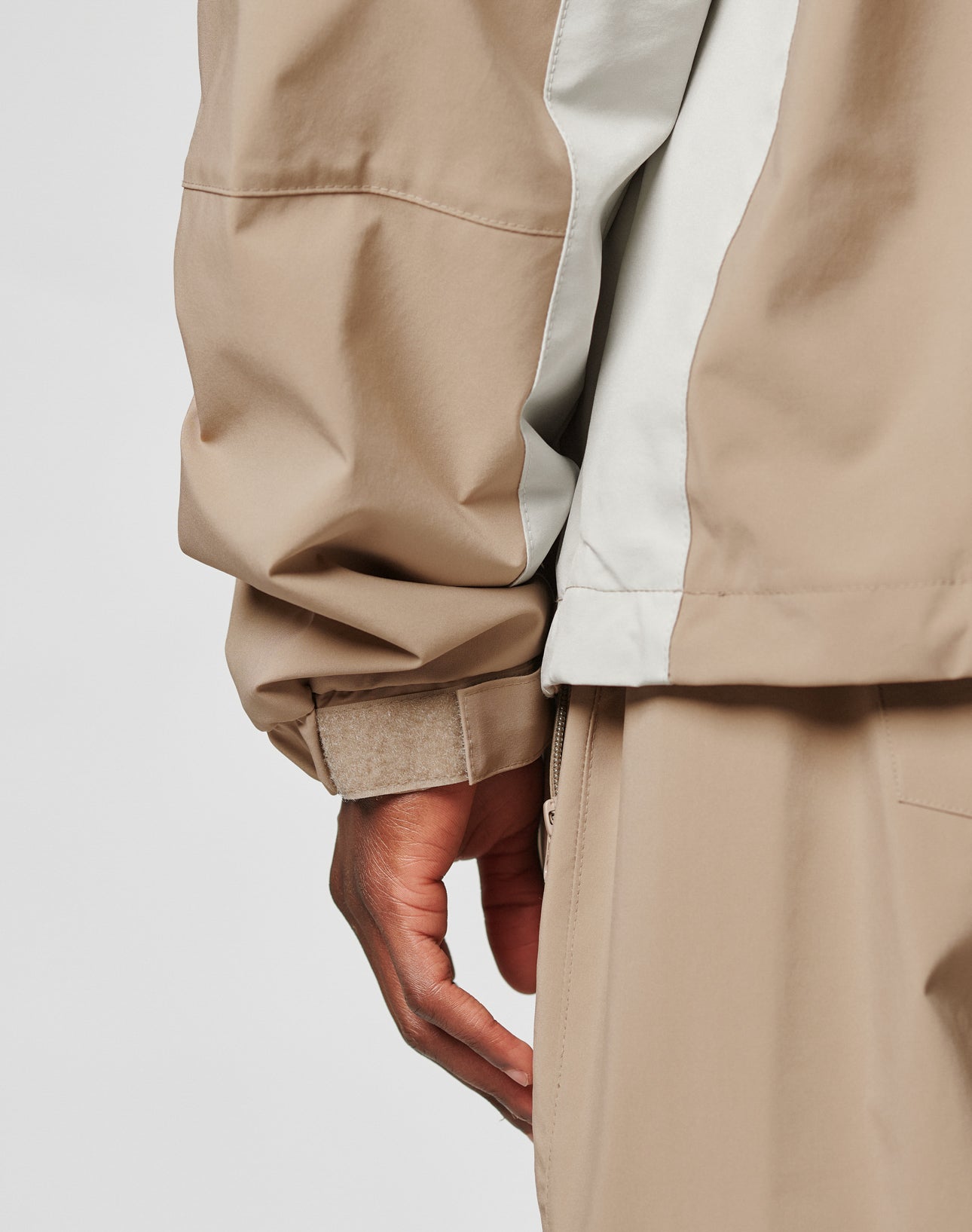 A close-up of a hand and arm wearing the LIVE FAST DIE YOUNG Basic Shell Jacket in an oversize fit light beige and off-white, with matching beige pants, highlighting the cuff, side details, and water-repellent zipper.