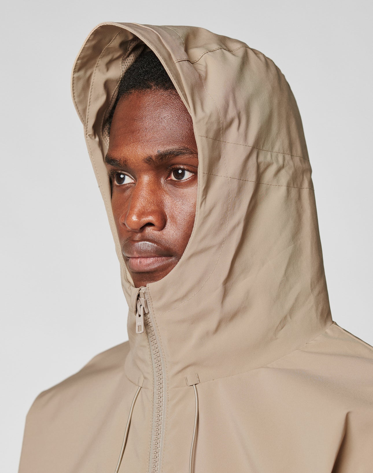 A person wears the LIVE FAST DIE YOUNG Basic Shell Jacket in beige with an oversized fit and hood up, featuring a wasserabweisender Reißverschluss, looking serious and gazing to the side against a plain light background.