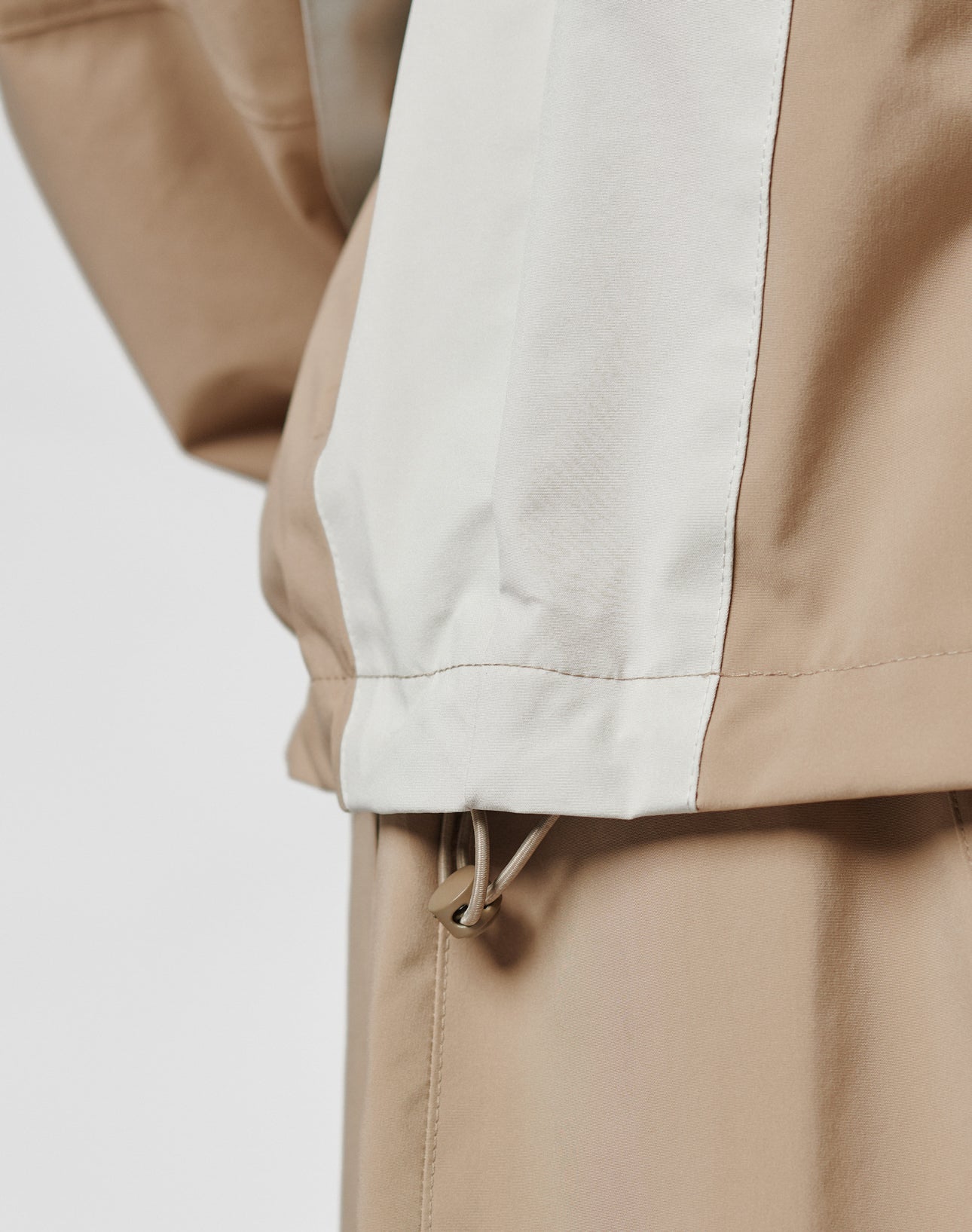 Close-up of the LIVE FAST DIE YOUNG Basic Shell Jacket in beige and light gray, featuring an oversize fit and drawstring hem, styled with matching beige pants. The fabric texture and drawstring detail pop against a neutral background.