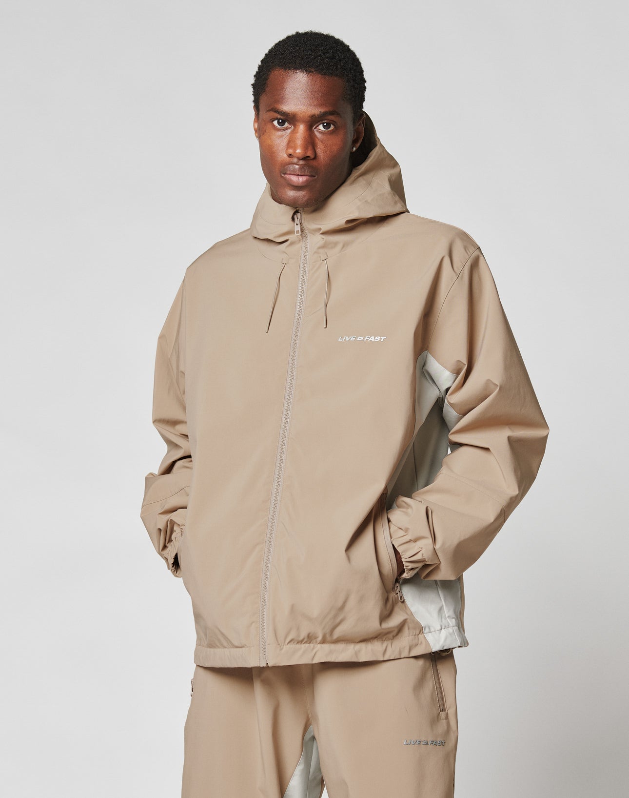 A man wears the LIVE FAST DIE YOUNG Basic Shell Jacket with reflektierender Druck and matching pants, standing hands in pockets and facing the camera against a plain white background.