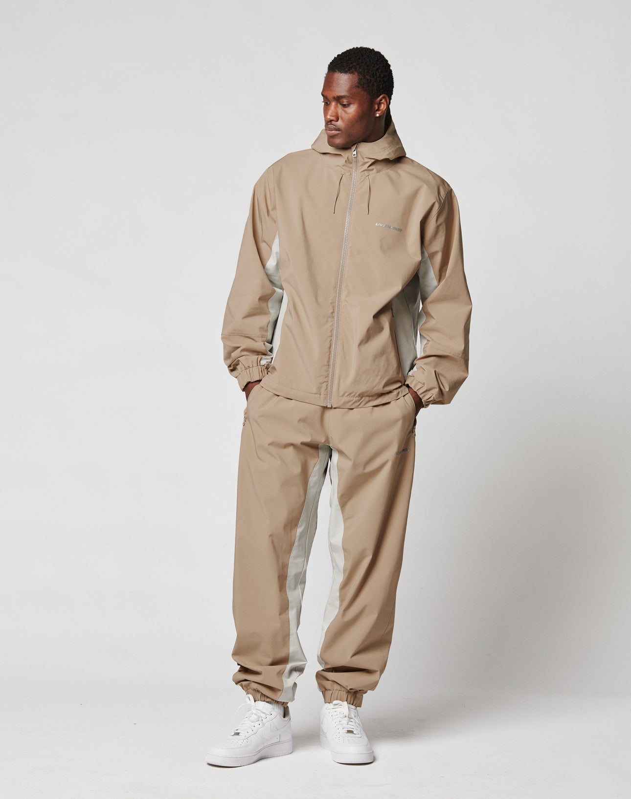 A man in a tan and gray LIVE FAST DIE YOUNG Basic Shell Jacket with a hood and oversize fit stands against a white background, looking to his left. The jacket features a wasserabweisender Reißverschluss and is paired with white sneakers.