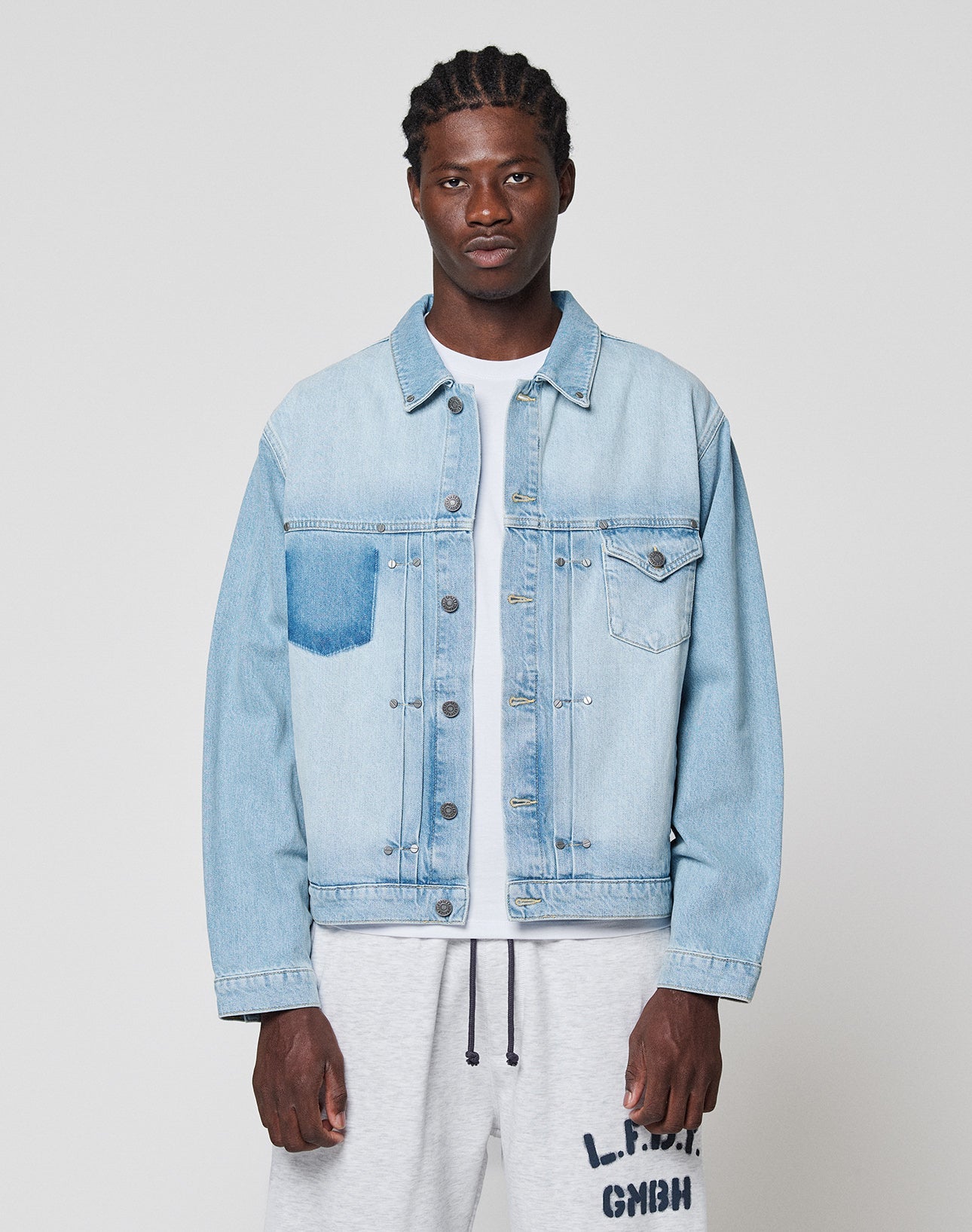 A man faces the camera wearing a LIVE FAST DIE YOUNG LFDY Trucker Jacket in light blue denim with one dark wash pocket, crafted from Baumwolle in Regular Fit, paired with a white T-shirt and light gray sweatpants with black text. Background is plain and light.