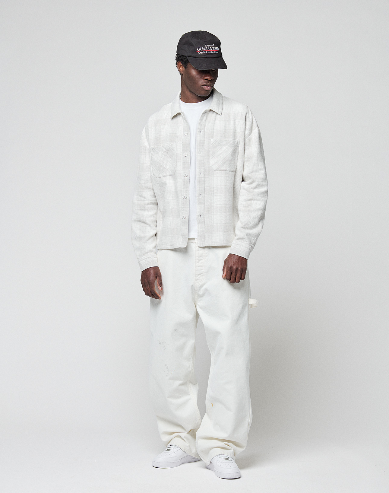 A person in a black cap, LIVE FAST DIE YOUNG Vintage Flannel with oversize fit, loose white pants, and white sneakers stands against a plain light-gray background.