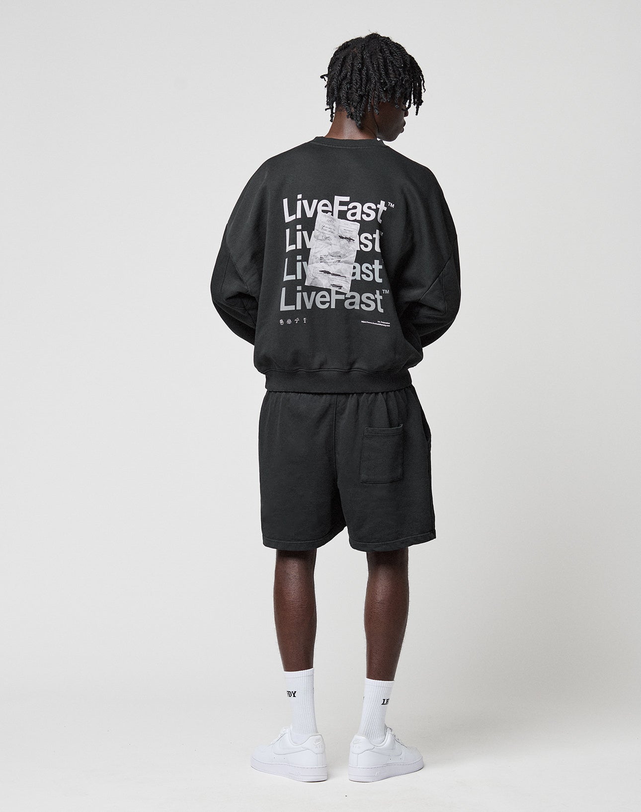 A person stands facing away, wearing a LIVE FAST DIE YOUNG Basic Evidence Sweater in oversize fit with front and back print, paired with black shorts, white socks, and white sneakers against a plain white background. Crafted from Baumwolle Polyester.