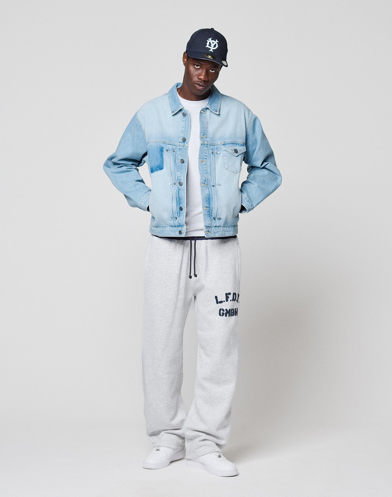 A man wearing the LIVE FAST DIE YOUNG LFDY Trucker Jacket, Regular Fit gray sweatpants with L.F.D.M. GmbH branding, a dark baseball cap, and white sneakers stands against a plain light background with his hands in his pockets.