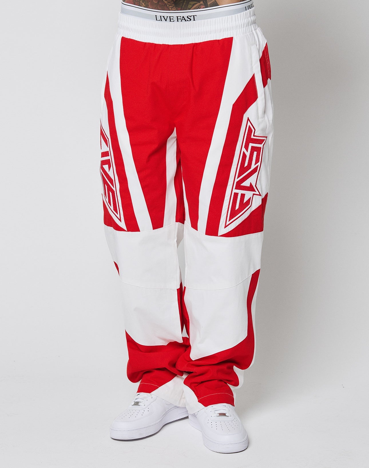 A person wears LFDY Cross Pants by LIVE FAST DIE YOUNG—red and white, bold geometric patterns, oversize fit, “FAST” on each leg, waistband reads “LIVE FAST”, 100% Baumwolle—paired with white sneakers against a plain white background.