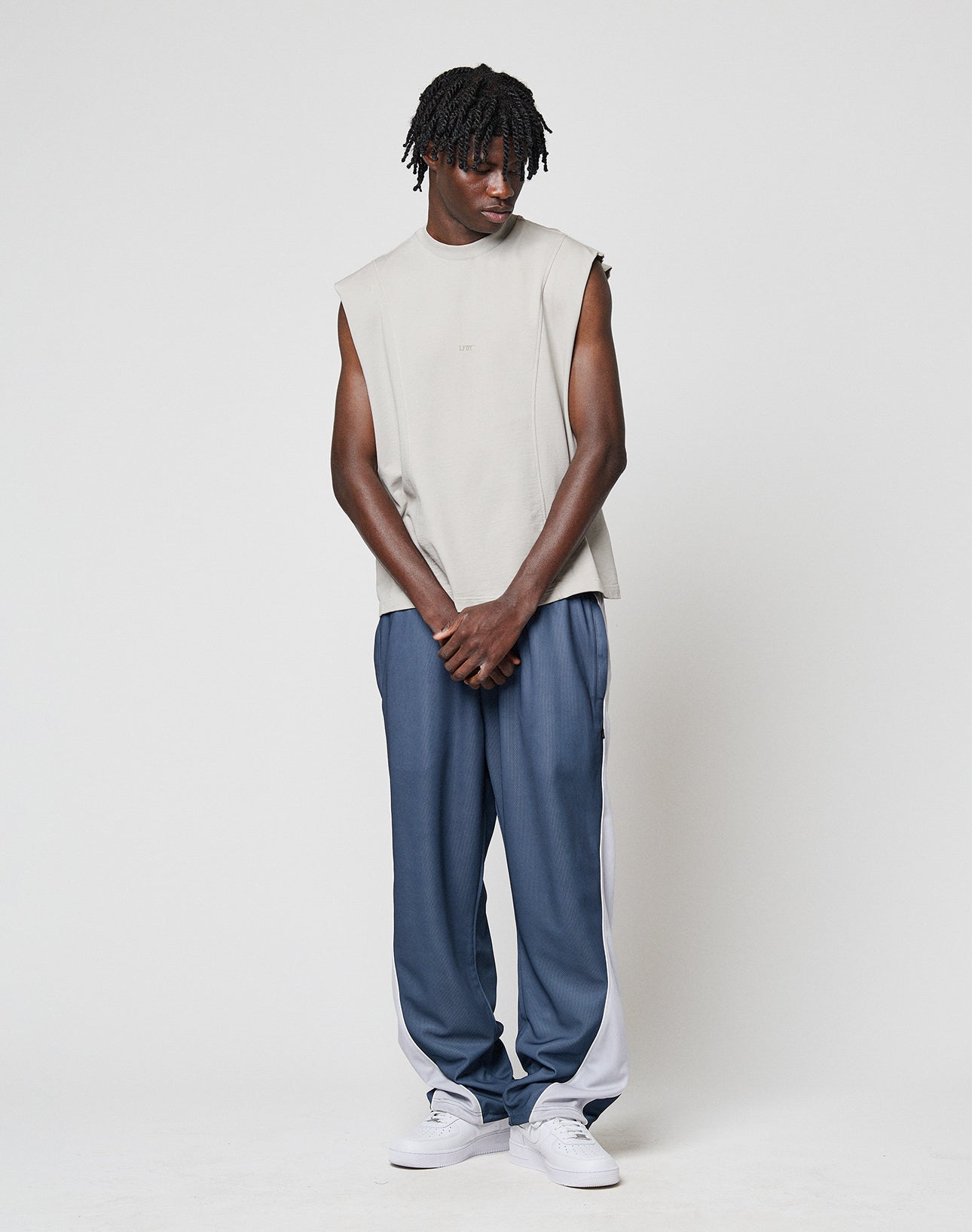A man with short dreadlocks wears the LIVE FAST DIE YOUNG Heavy Muscle Tank in light grey, styled in an oversize fit, paired with loose blue track pants with white side panels and white sneakers, standing against a plain white background looking down.