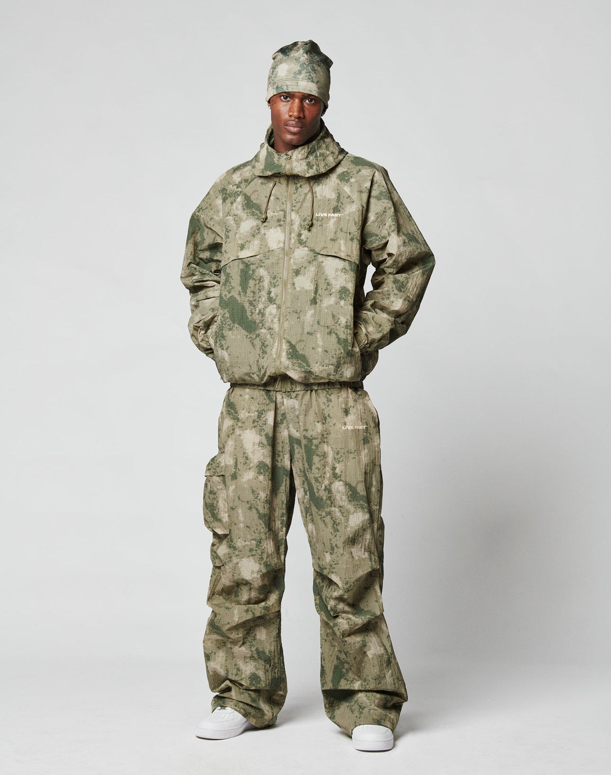 A person stands against a plain background wearing the LIVE FAST DIE YOUNG Live Fast Nylon Jacket in an oversize green camouflage style with Kapuze mit Tunnelzug, matching pants, and white sneakers. Their hands rest in the jacket’s Reißverschluss Taschen for a relaxed look.