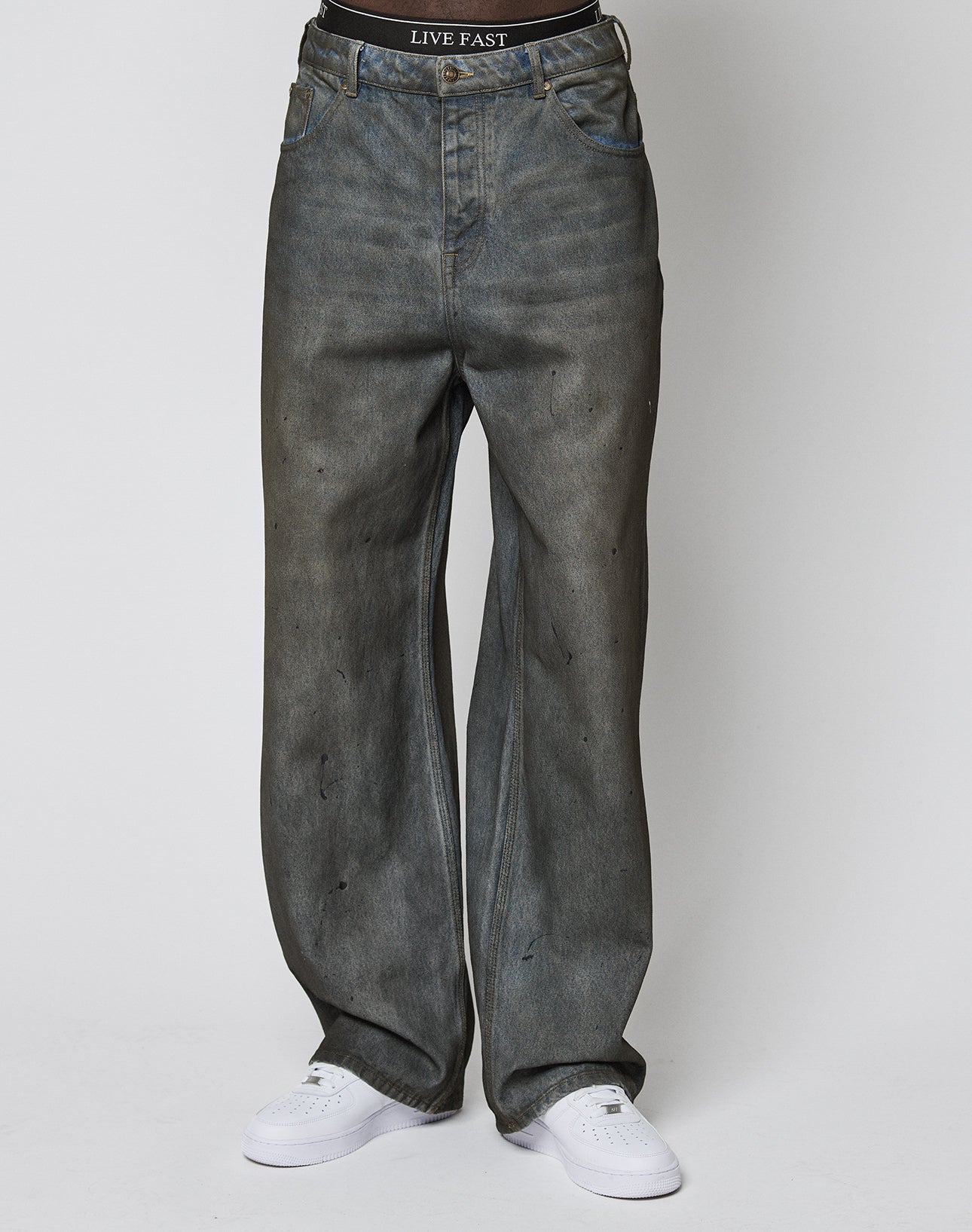 A person wearing LIVE FAST DIE YOUNGs LFDY Coated Baggy Denim—baggy fit, faded gray with black spots—and a black LIVE FAST waistband stands against a plain white background in white sneakers.