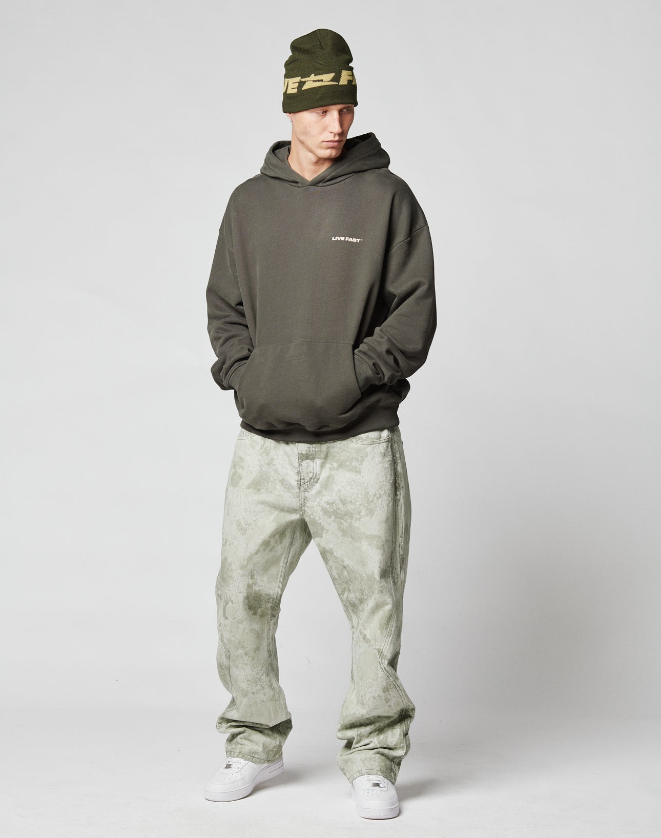 A person wearing the Live Fast Plain Hooded by LIVE FAST DIE YOUNG, a dark green oversized hoodie, paired with a green beanie, light green patterned baggy jeans, and white sneakers stands with hands in pockets against a plain white background.