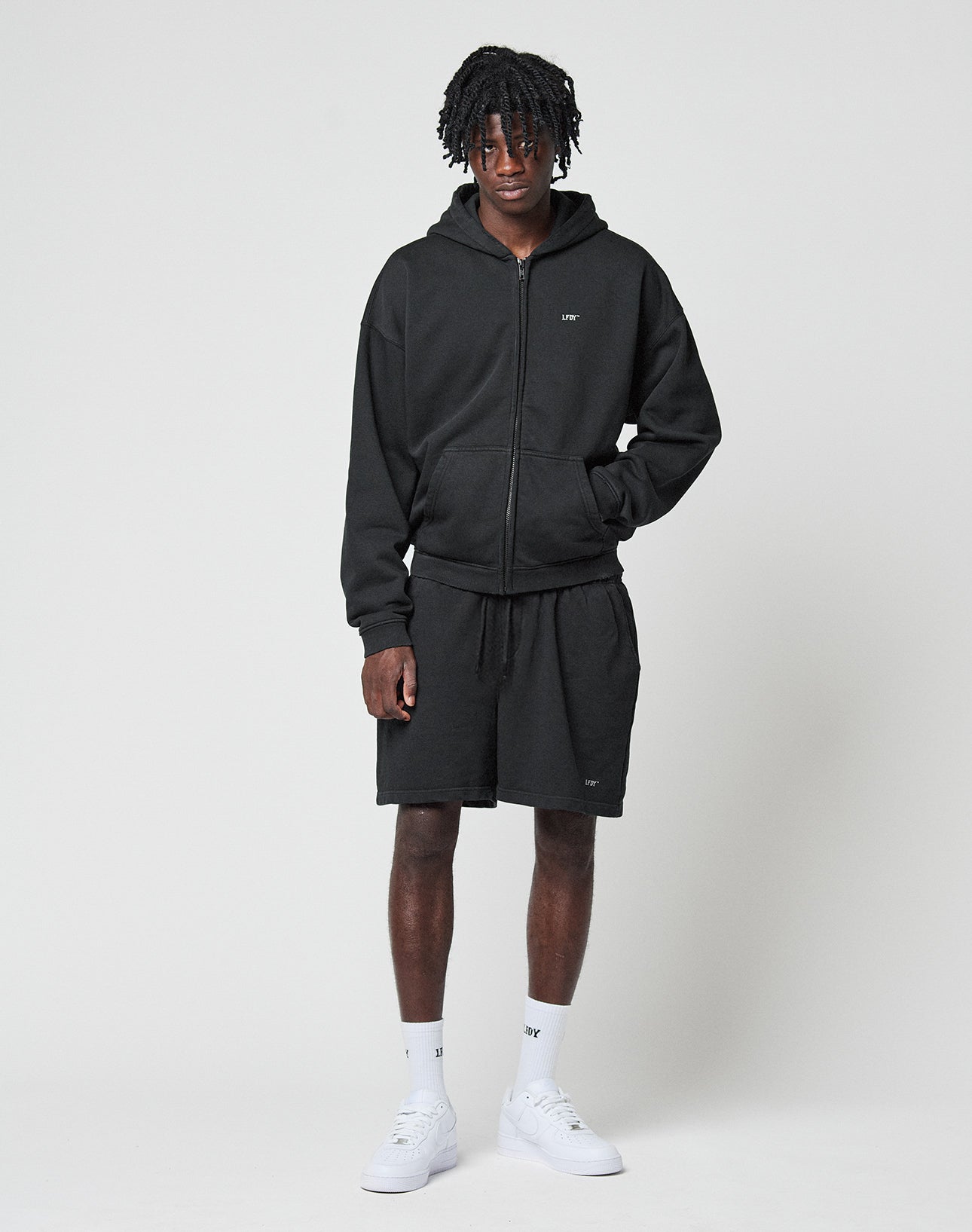 A person stands against a plain white background wearing a black LIVE FAST DIE YOUNG Basic Hooded Zip, matching black shorts, white crew socks, and white sneakers. One hand is in their pocket and they look forward with a slight smile.