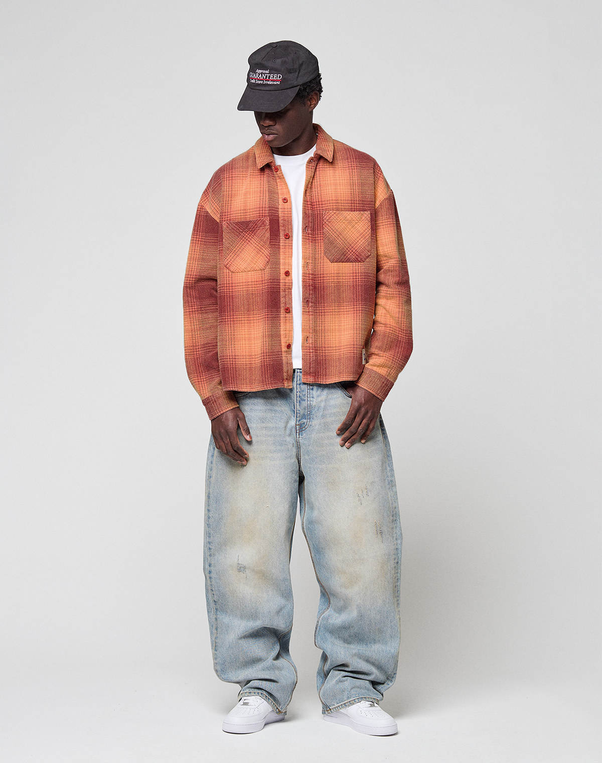A person in a dark cap, LIVE FAST DIE YOUNG Vintage Flannel in orange plaid over a white tee, baggy light blue jeans with vintage wash, and white sneakers stands against a plain light gray background, looking downwards.