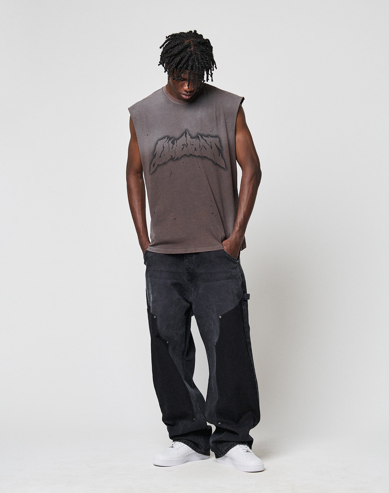 A young man with short dreadlocks wears the LFDY Flames Muscle Shirt by LIVE FAST DIE YOUNG—a sleeveless, faded gray top with handgefertigtem Sprayeffekt—paired with loose black jeans and white sneakers, standing against a plain background.
