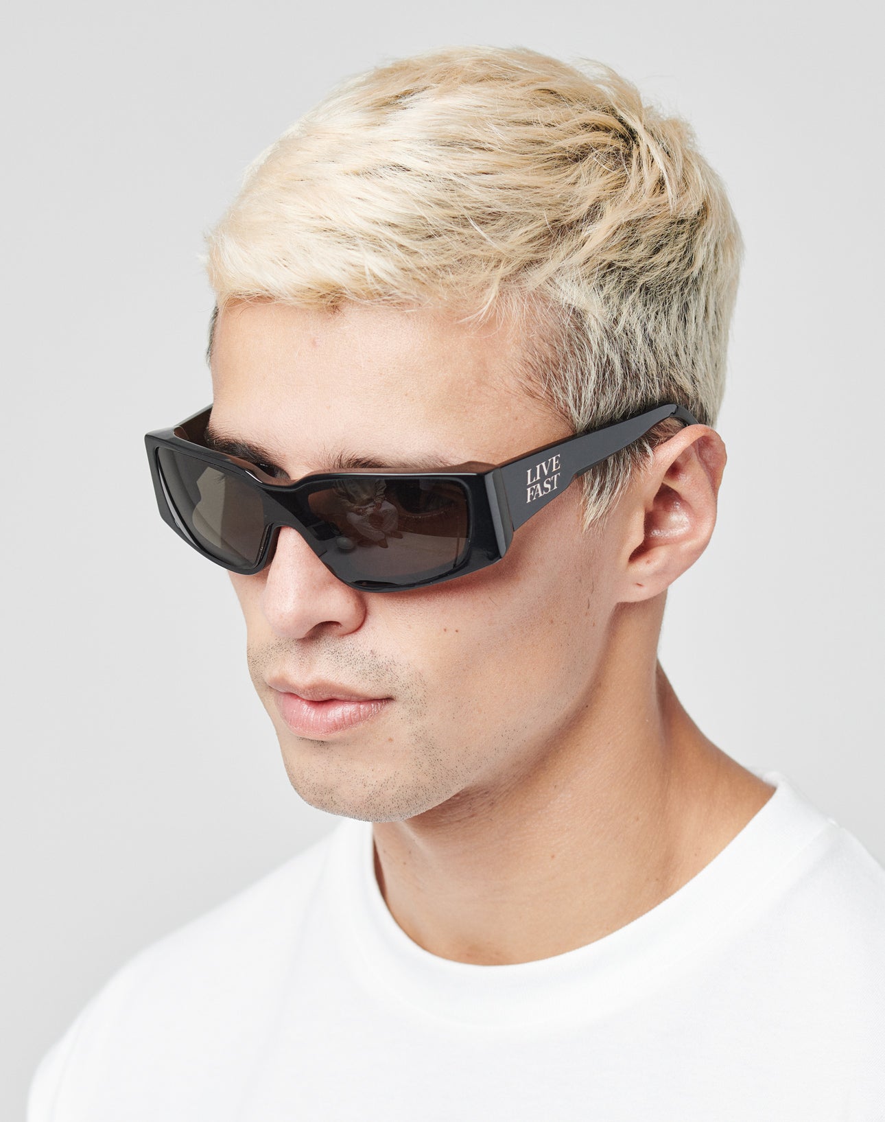 A man with short, bleached blond hair wears LIVE FAST DIE YOUNGs Live Fast Curved Shades—black rectangular Mazzuchelli Acetate sunglasses with 400 UV protection and LIVE FAST on the side—paired with a plain white shirt against a light background.