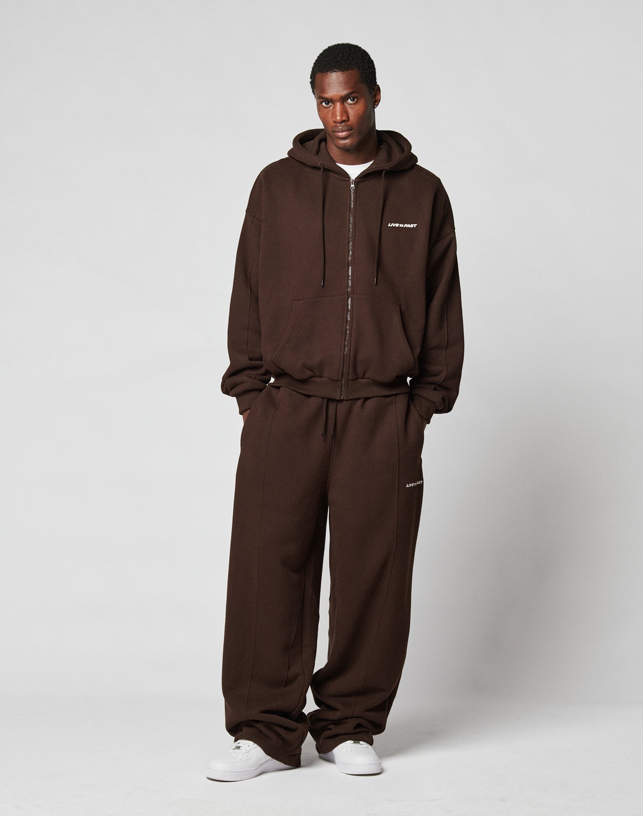 A person stands against a plain light background, wearing LIVE FAST DIE YOUNG’s LFDY Pleated Jogger and a matching wide fit brown zip-up hoodie with Baumwolle elastik-bund and a small white chest logo, paired with white sneakers.
