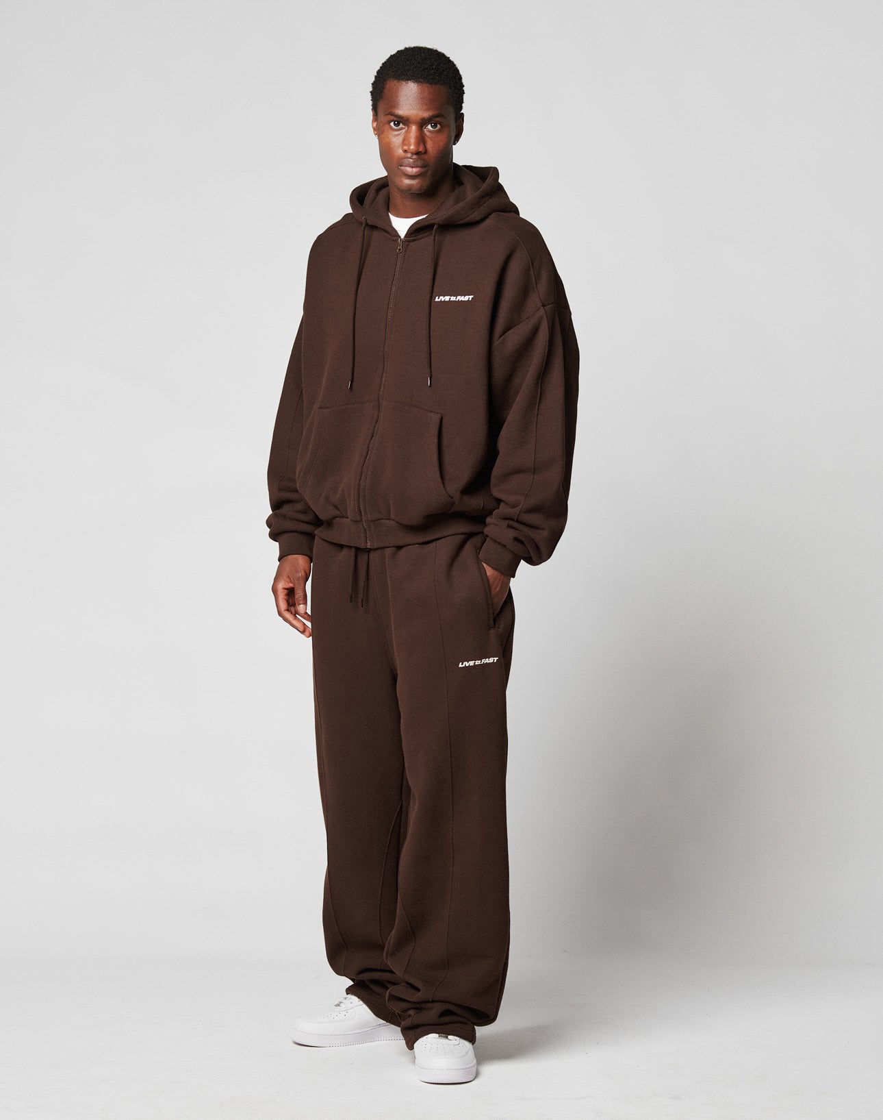 A man stands against a plain background wearing the LFDY Pleated Hooded Zip from LIVE FAST DIE YOUNG in dark brown with oversized fit, hood, white text details, matching sweatpants, and white sneakers.