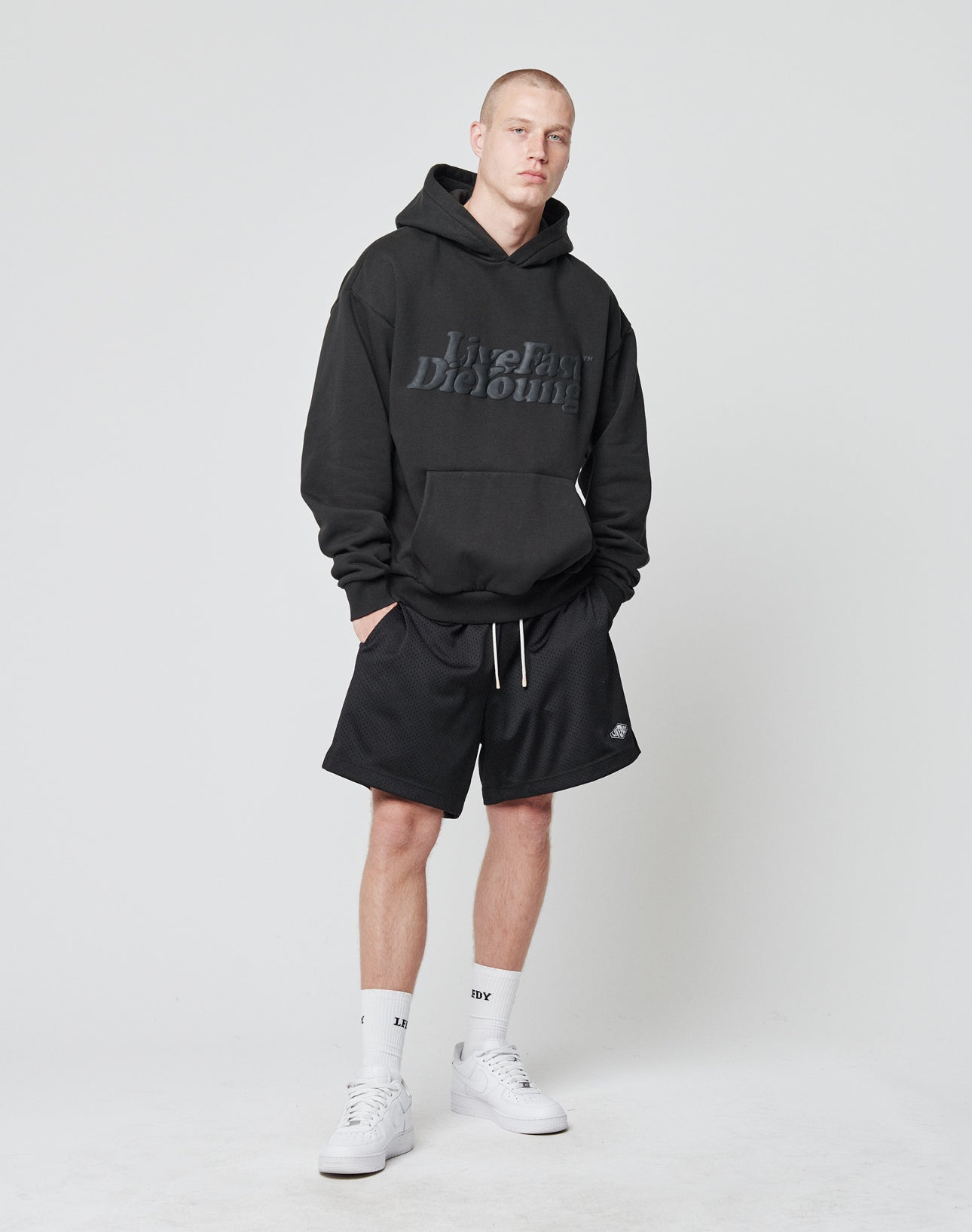 A man with short hair stands against a plain background wearing a black LIVE FAST DIE YOUNG hoodie, LIVE FAST DIE YOUNG Basic Mesh Shorts with elastic waistband, white crew socks, and white sneakers. His hands are in his hoodie pocket.
