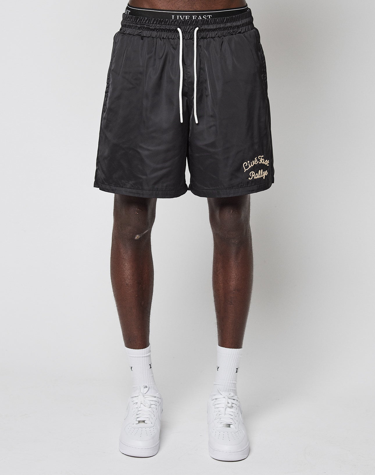 A person wears black LFDY Retro Satin Shorts by LIVE FAST DIE YOUNG, featuring a yellow chainstitch logo, with white socks and sneakers, standing against a plain white background.