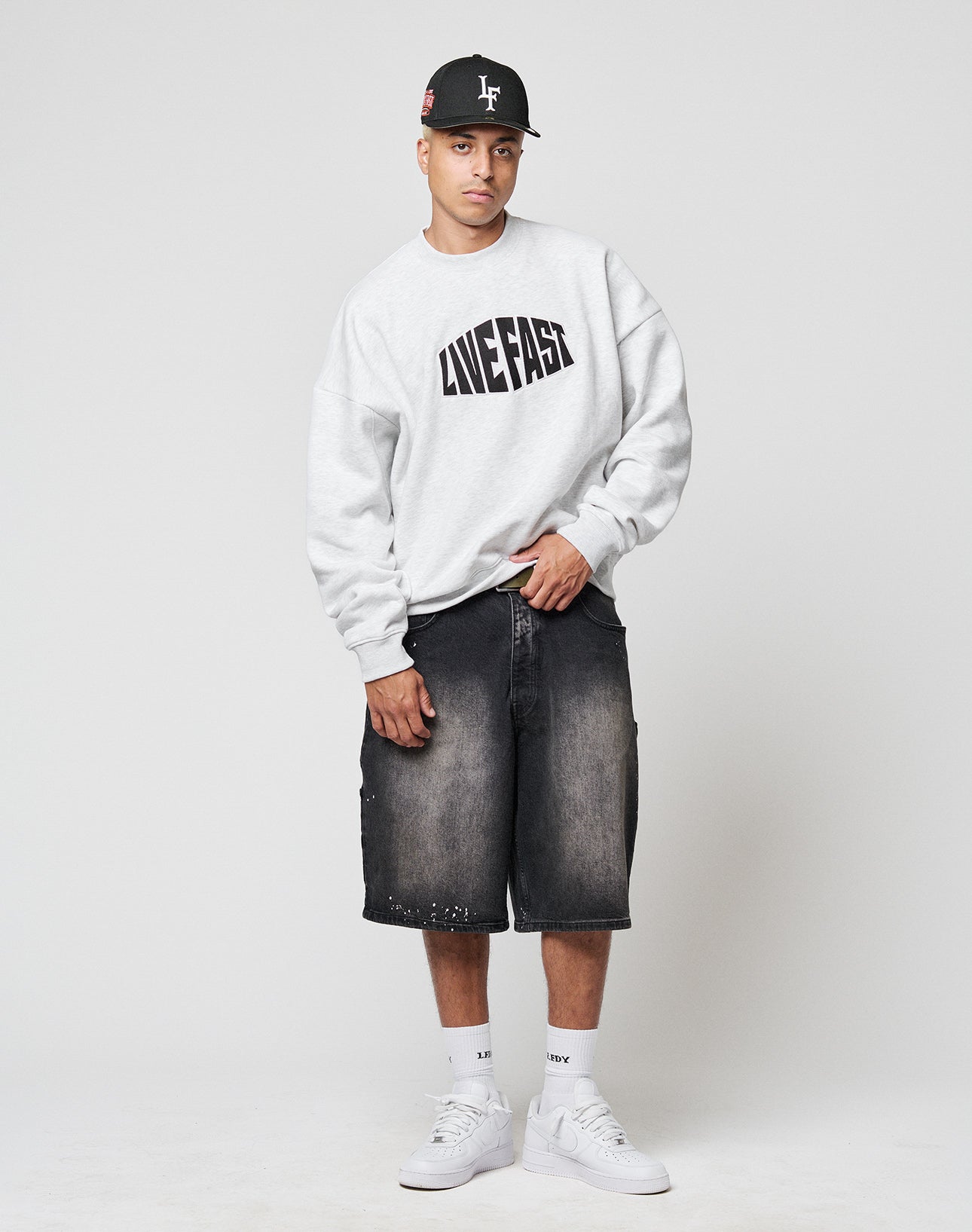 A man in an oversized gray LFDY Emblem Sweater by LIVE FAST DIE YOUNG, featuring a subtle logo embroidery, black cap, black denim shorts, white socks, and white sneakers stands against a plain white background.