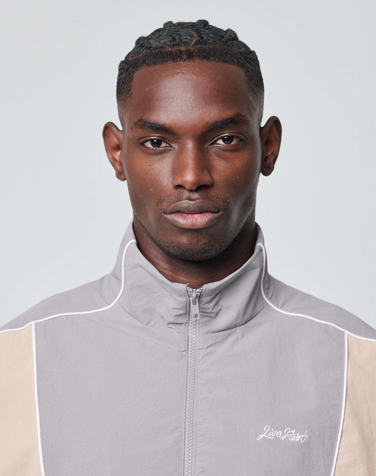 A man with short, styled hair wears a light gray and beige Basic Nylon Trackjacket from LIVE FAST DIE YOUNG, looking directly at the camera with a neutral expression against a plain background.