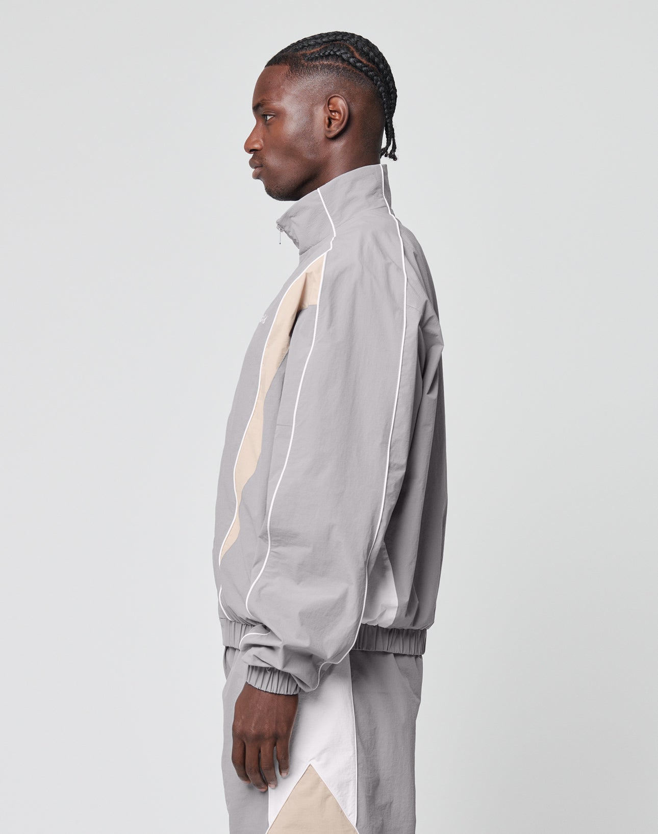 A person in profile wears a light gray LIVE FAST DIE YOUNG Basic Nylon Trackjacket with a high collar, beige and white accents, Logo-Stick detail, and matching pants with beige side design. The background is plain and light-colored.