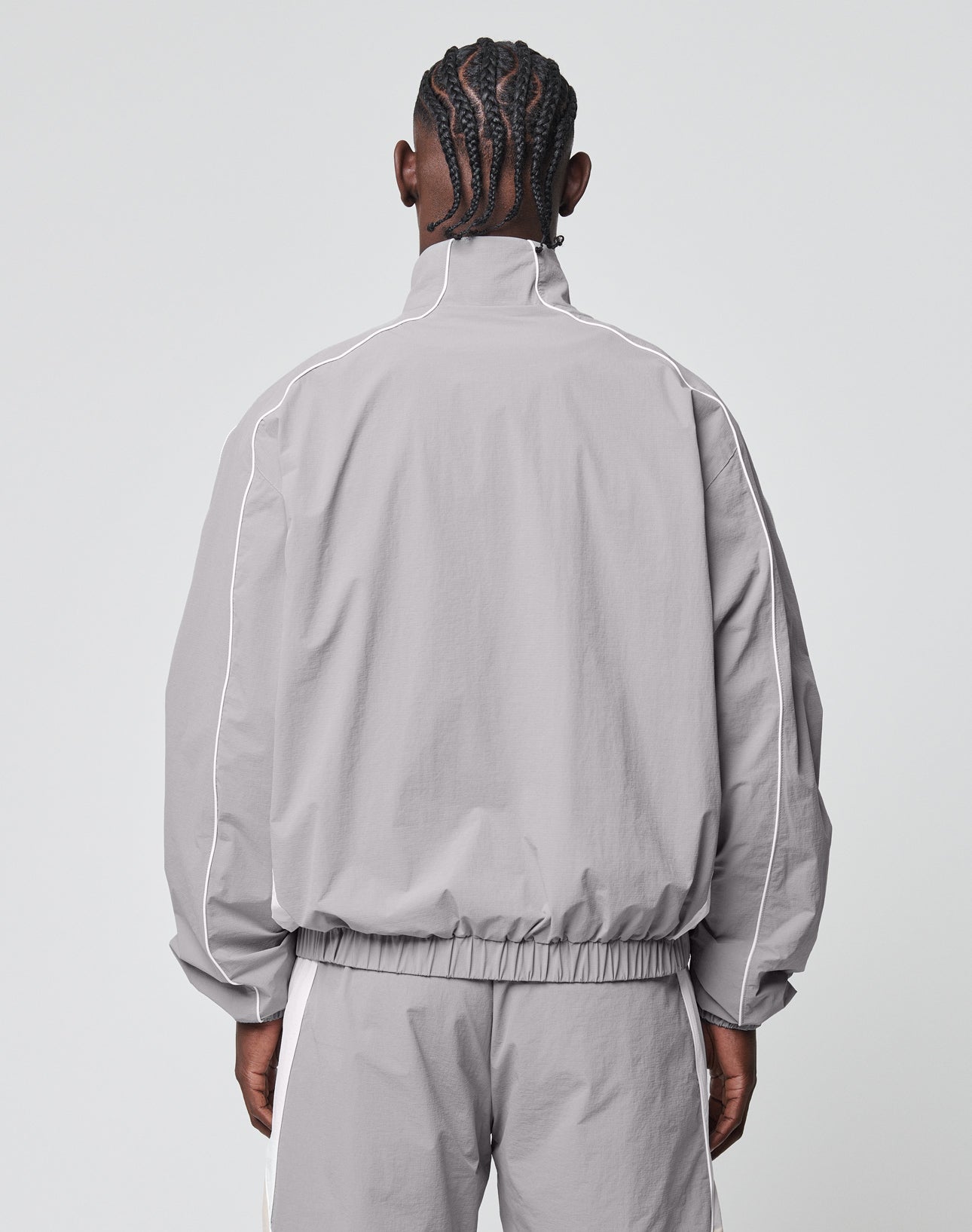 A person with braided hair is shown from the back, wearing the LIVE FAST DIE YOUNG Basic Nylon Trackjacket and matching pants in light gray with white piping, set against a plain light background.