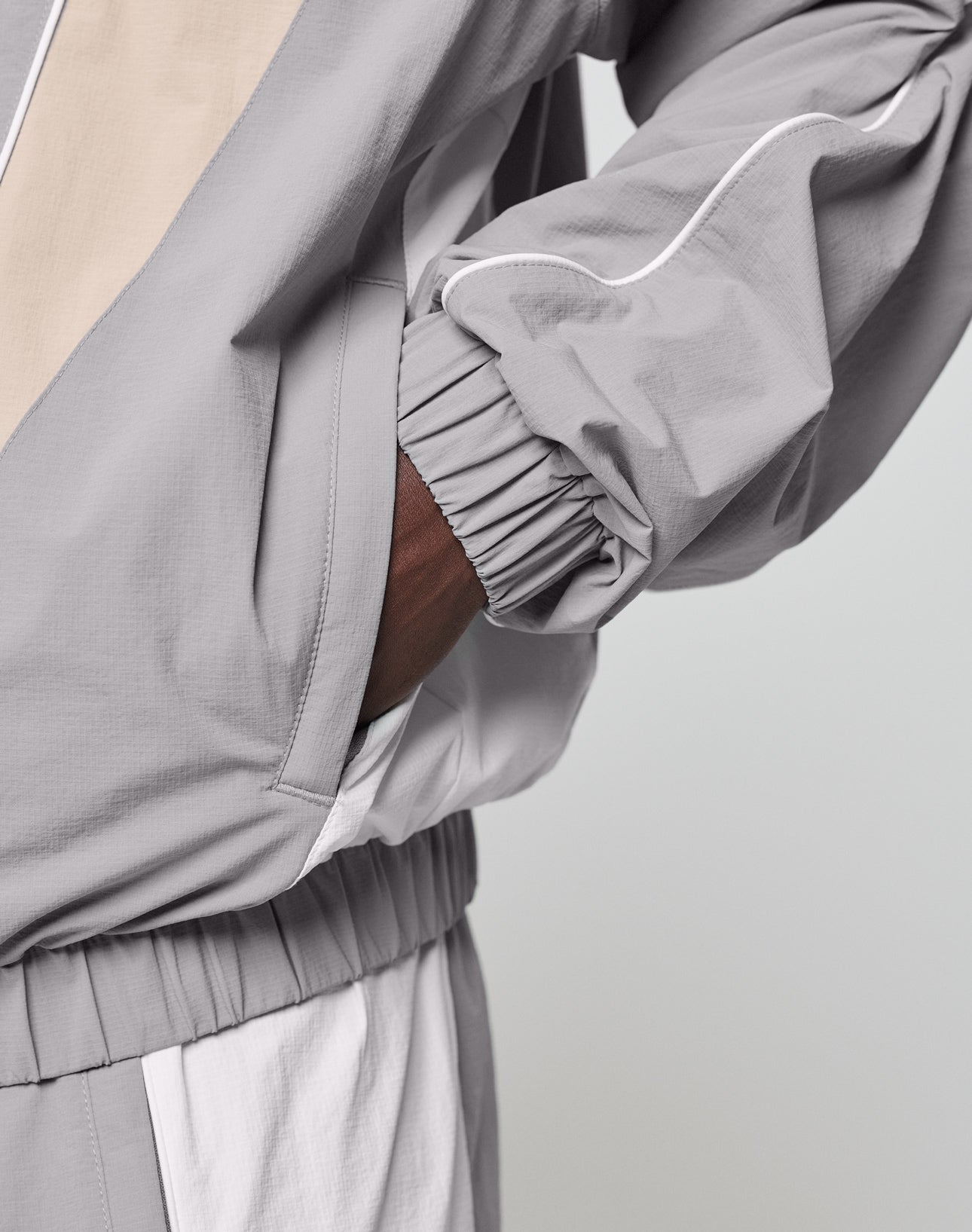 A person wears the LIVE FAST DIE YOUNG Basic Nylon Trackjacket in light gray and beige with matching pants, featuring a regular fit, elastic cuffs and waistband, color-block design, and hand in the jacket pocket.