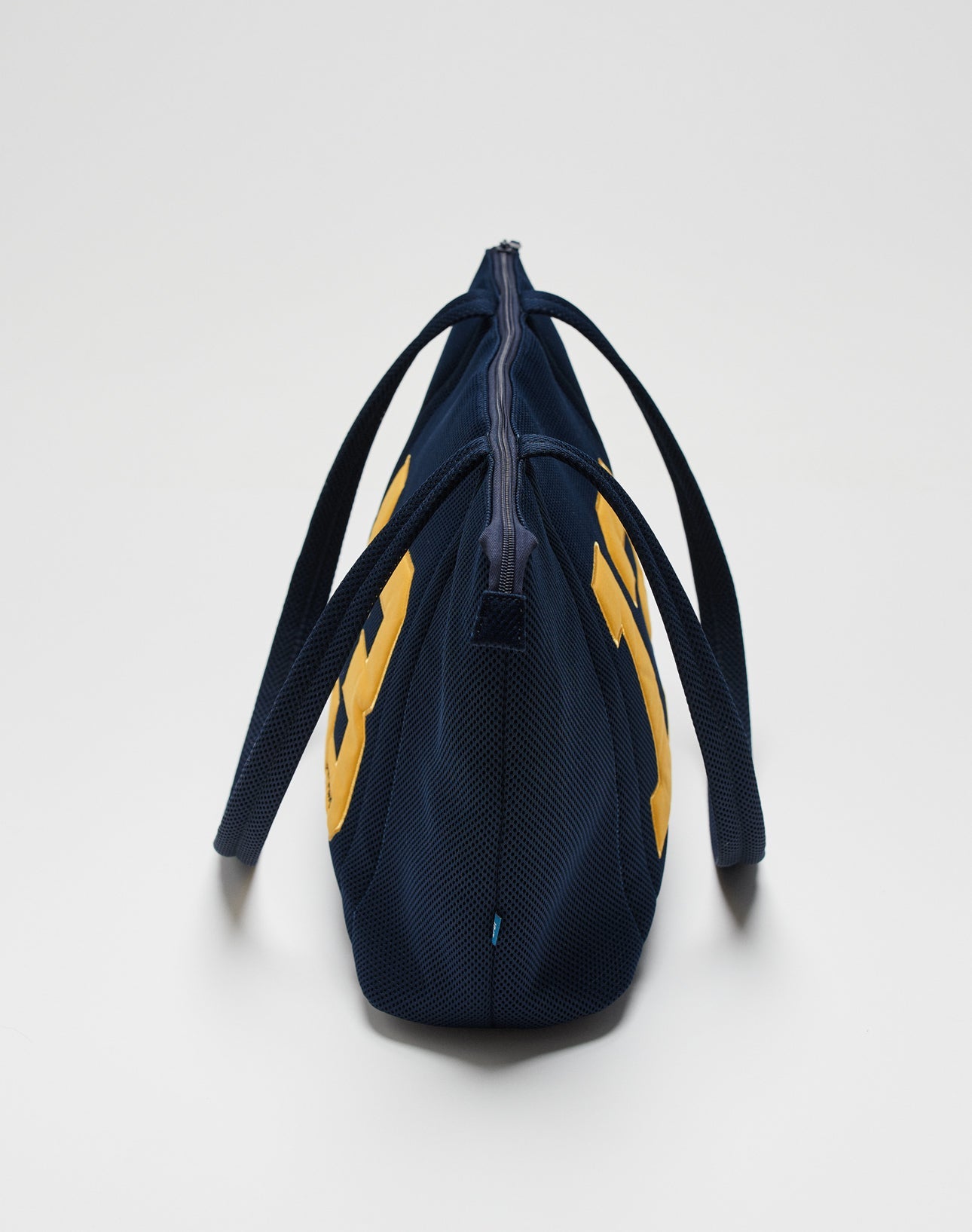The LFDY Mesh Beach Bag by LIVE FAST DIE YOUNG is shown from the side with two handles, navy blue color with yellow accents, a zippered top closure, and a plain white background.