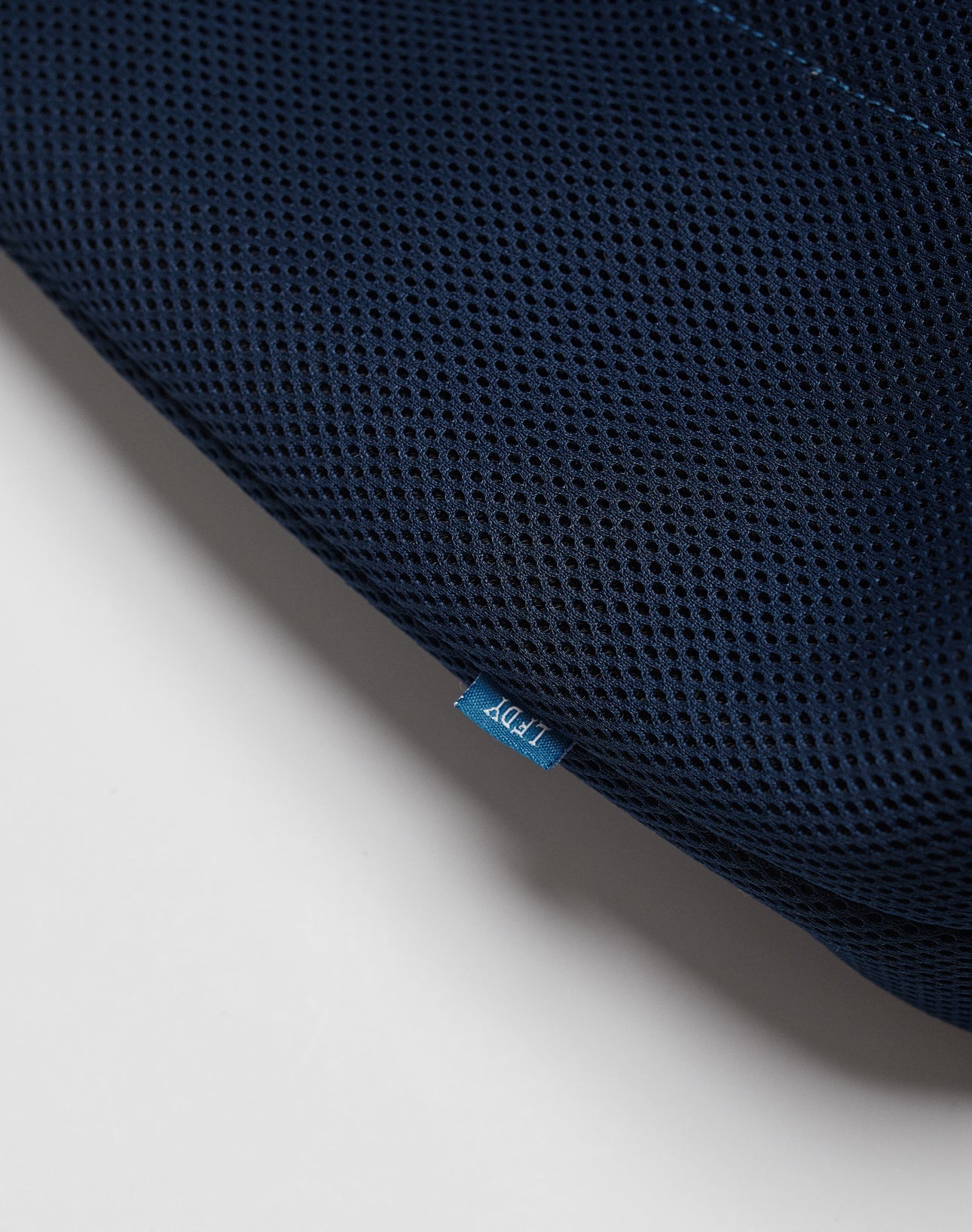 Close-up of dark blue textured mesh fabric from the LFDY Mesh Beach Bag by LIVE FAST DIE YOUNG, with a small blue tag showing white text and part of the bag visible against a plain white background.