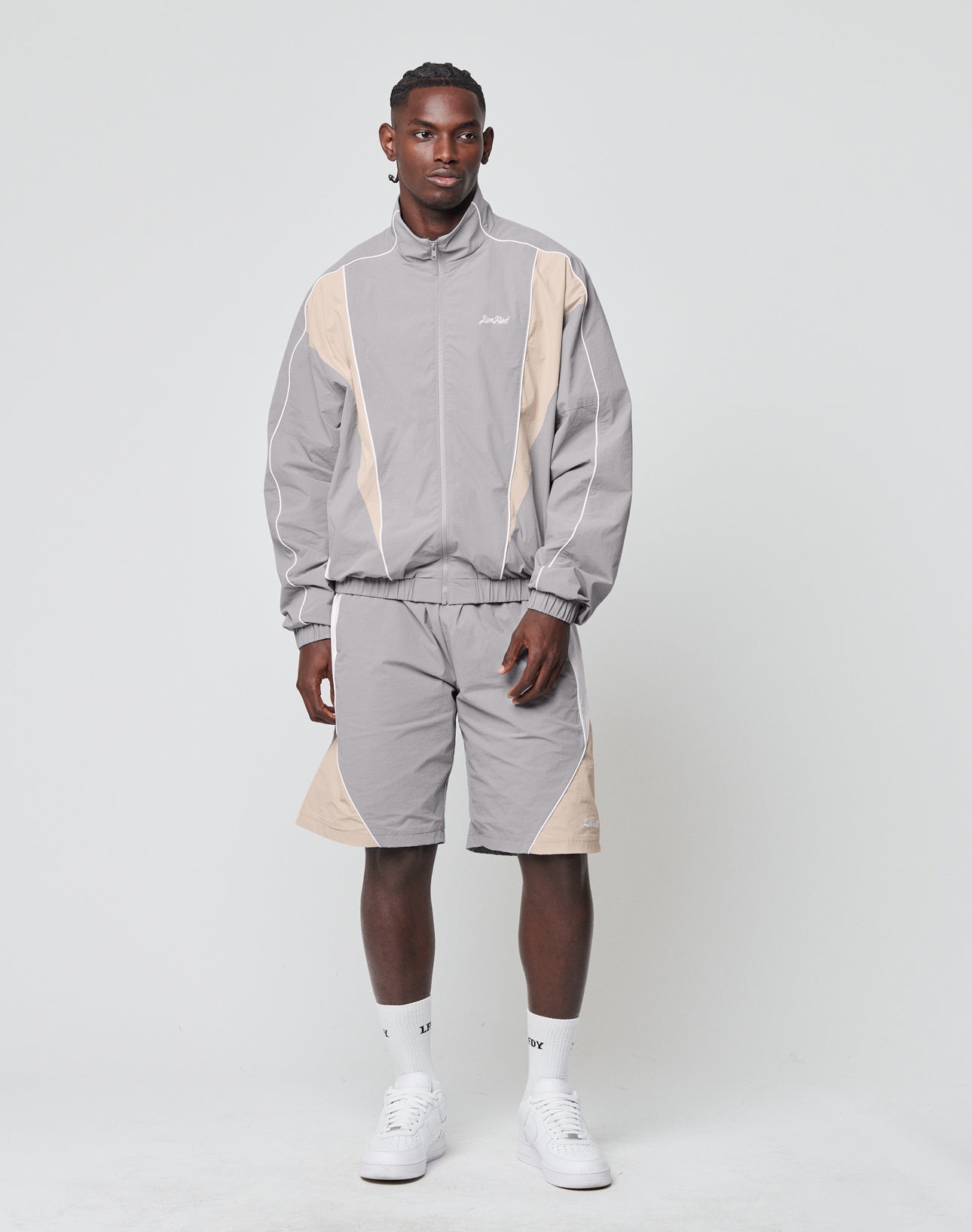 A man stands against a white background wearing LIVE FAST DIE YOUNG’s Basic Nylon Trackshorts with a matching zip-up jacket, both in light gray and beige nylon-lycra, paired with white socks and sneakers.