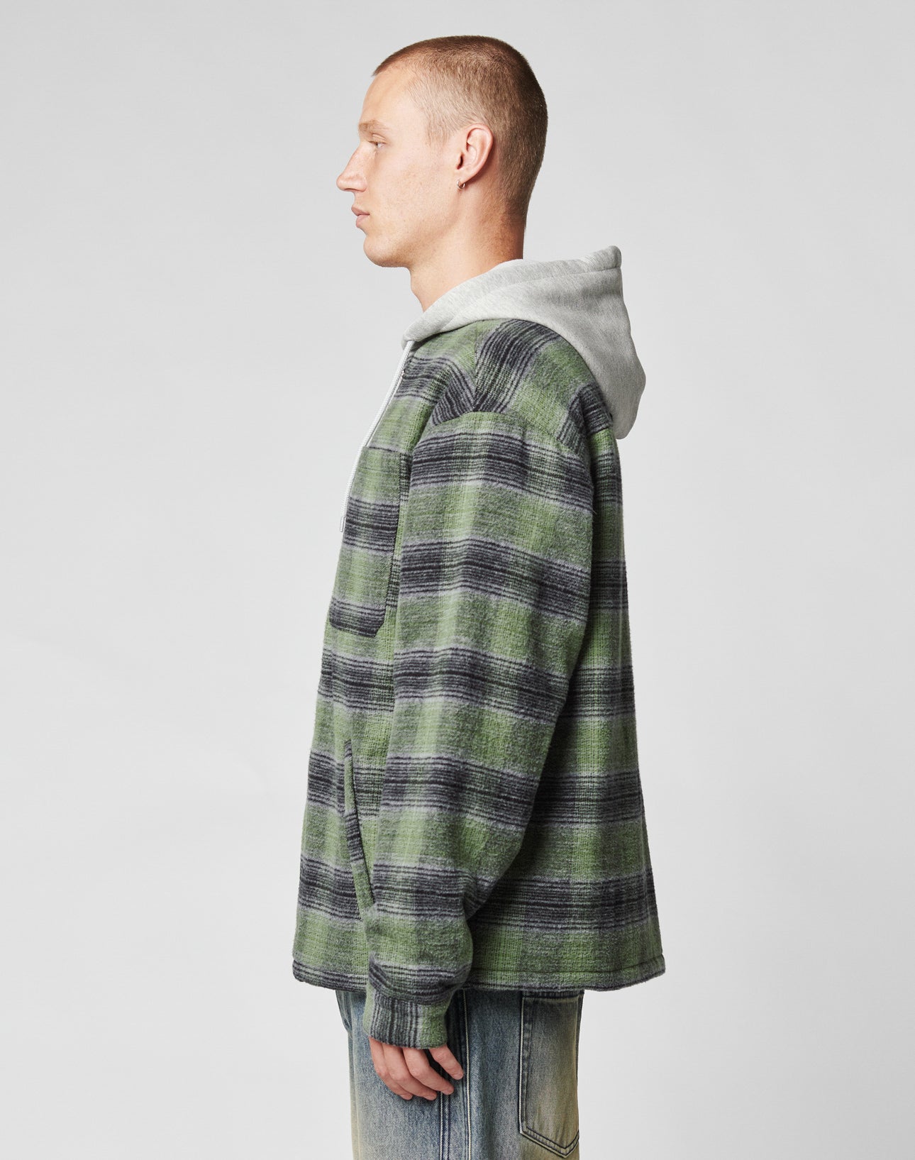A person stands in profile against a plain background, wearing the LIVE FAST DIE YOUNG Vintage Padded Hooded Flannel in green and black plaid, layered over a gray waffle-knit hoodie and paired with light blue jeans.