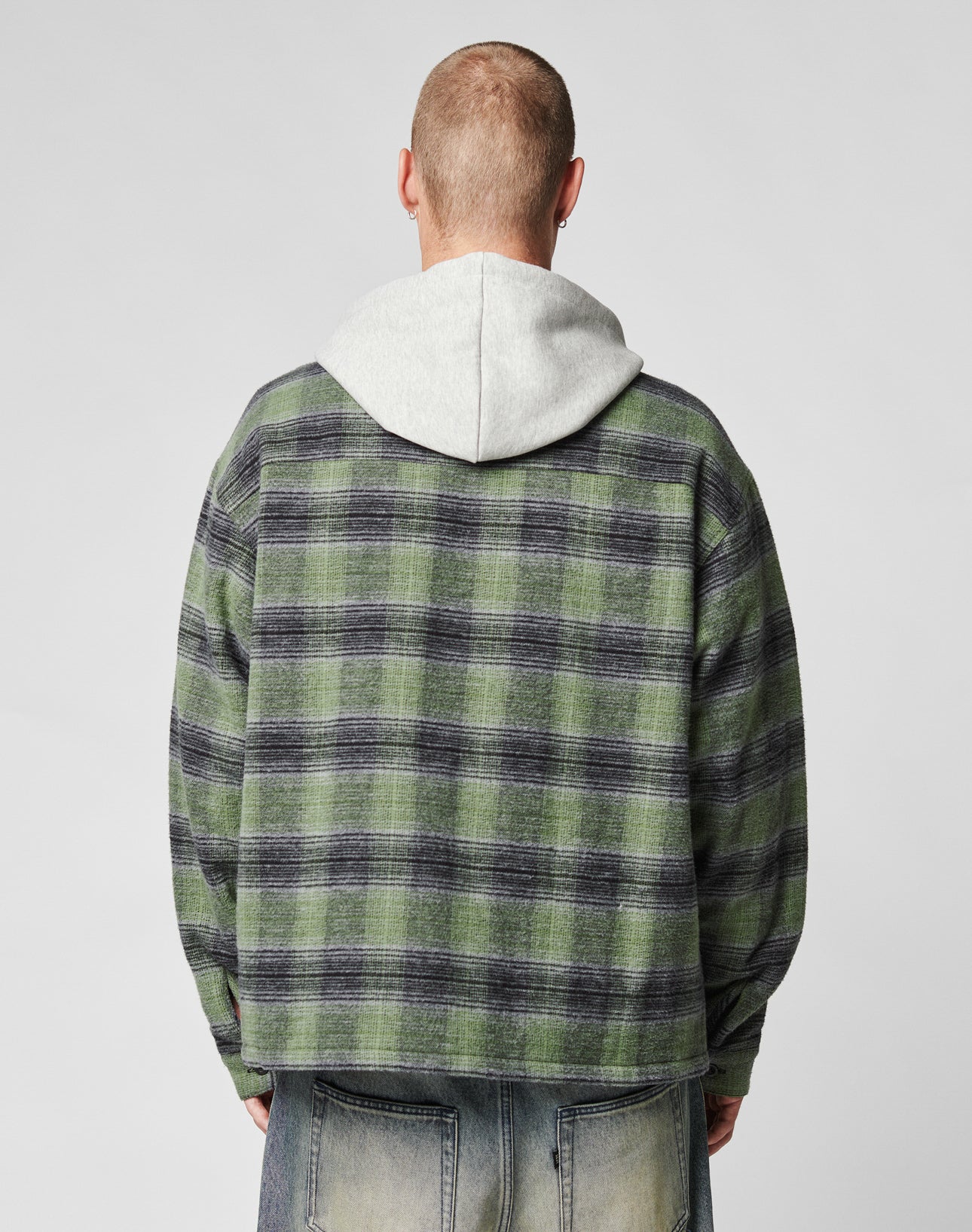 A person with short hair, seen from behind, wears a green and black LIVE FAST DIE YOUNG Vintage Padded Hooded Flannel over a light gray waffle-lined hoodie and faded blue jeans against a plain, light background.