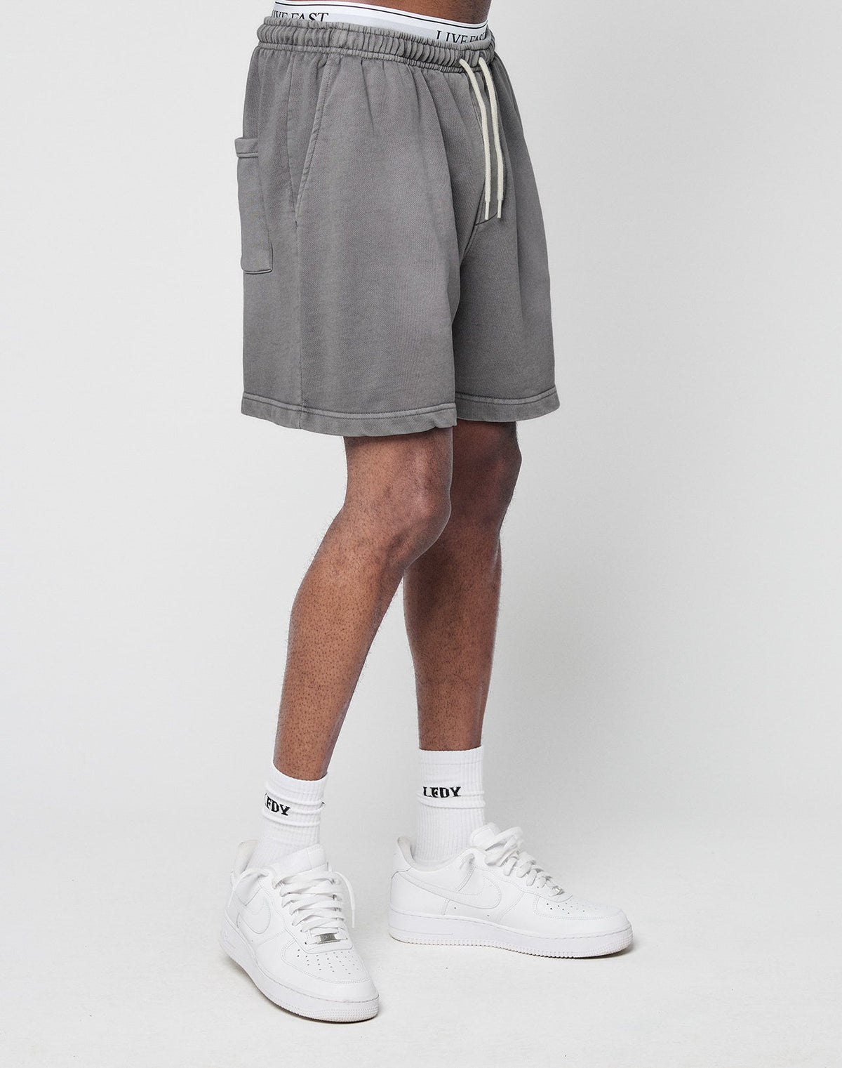 A person wearing LIVE FAST DIE YOUNG Basic Sweat Shorts in gray with an elastic waistband, white socks featuring a LUCKY logo print, and white sneakers stands against a plain white background.