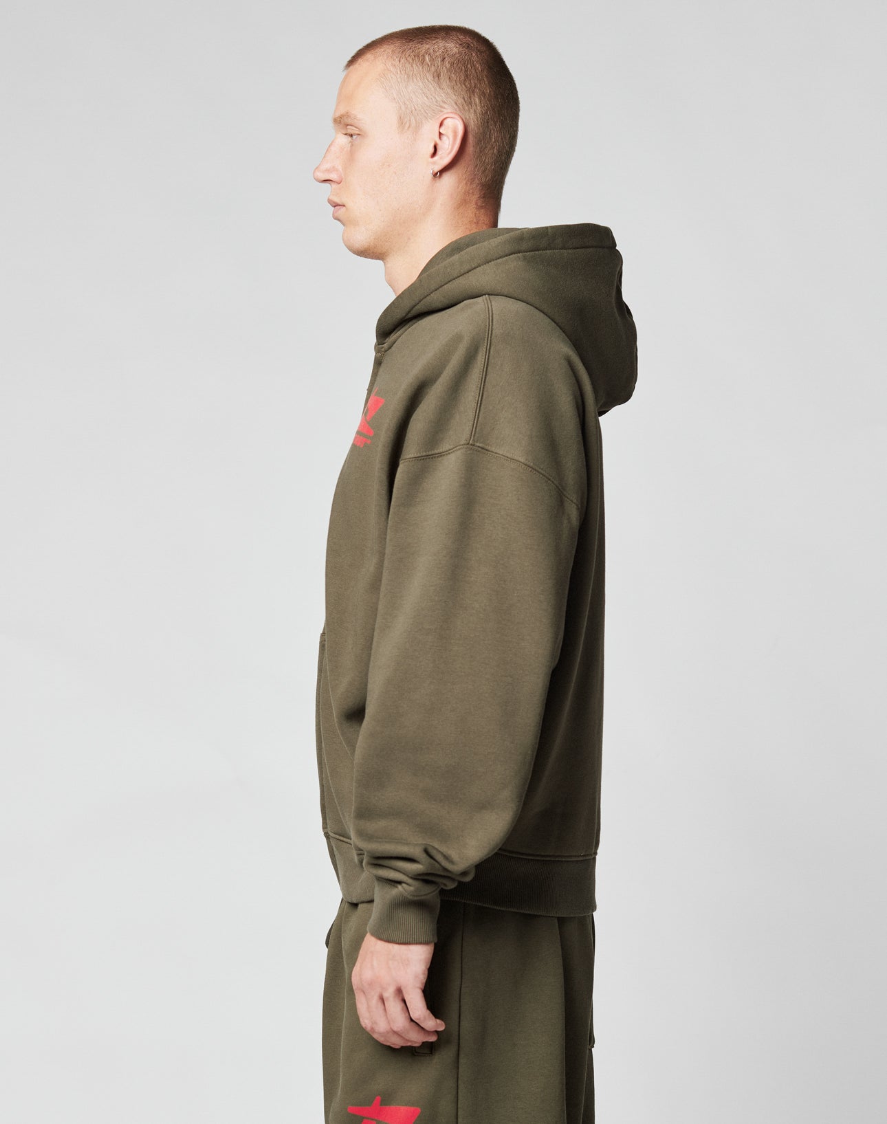 A man with short hair stands in profile, wearing the LIVE FAST DIE YOUNG Live Fast Sweatsuit Jacket in olive green Oversize Fit and matching sweatpants with red front print, set against a plain light gray background.
