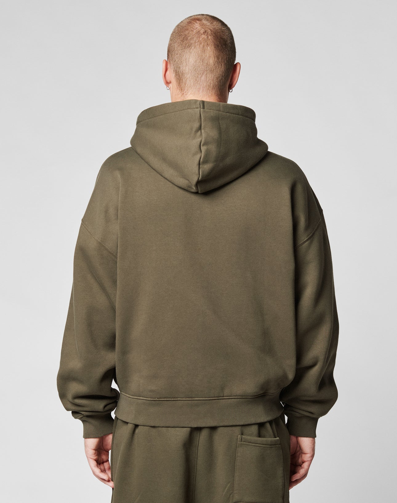A person with a shaved head is shown from behind in a LIVE FAST DIE YOUNG Live Fast Sweatsuit Jacket, oversize fit olive green, hood up, against a plain light gray background.