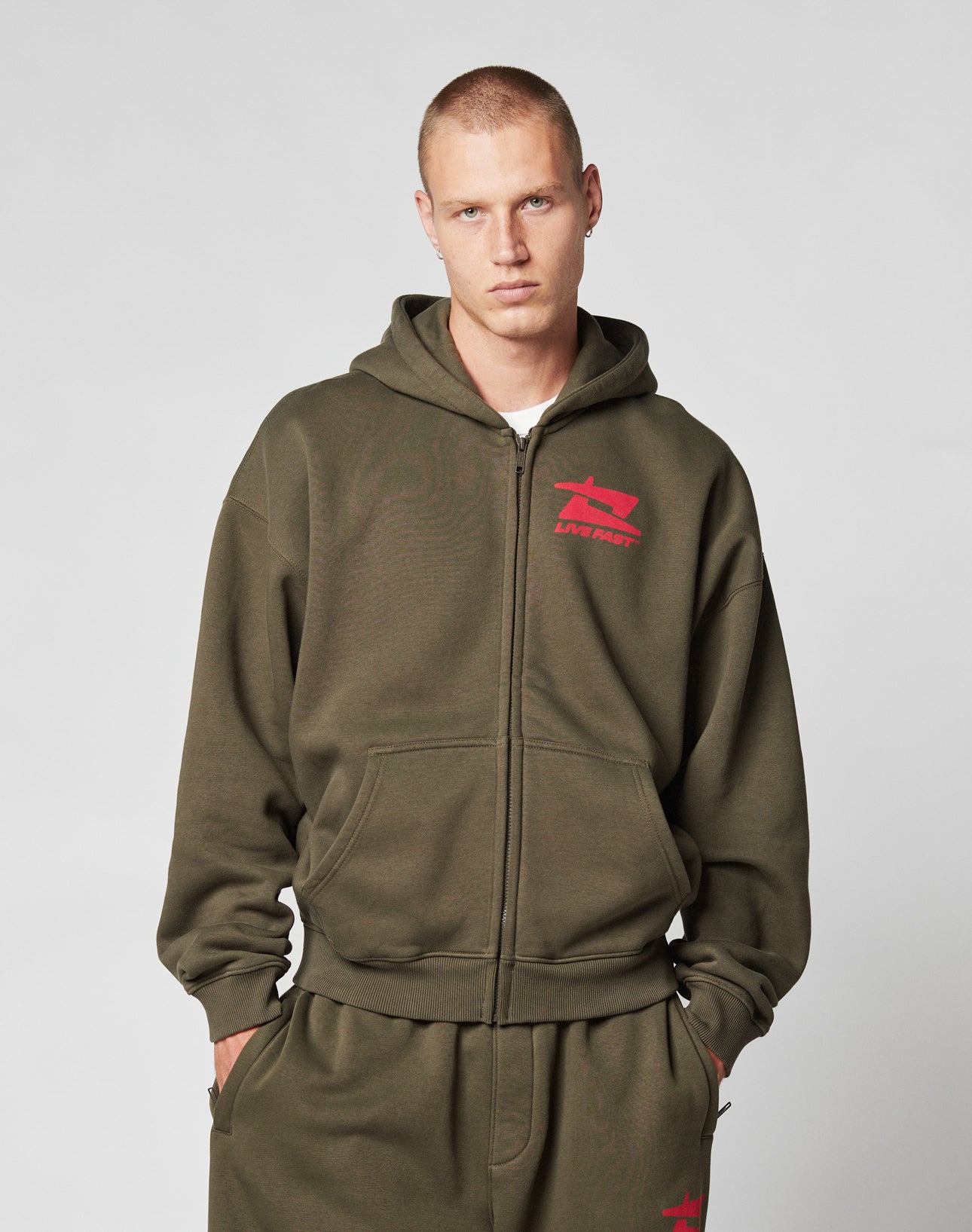 A young man with short hair wears the LIVE FAST DIE YOUNG Live Fast Sweatsuit Jacket in olive green, styled oversized with bold red front-print logos, and matching sweatpants, standing hands-in-pockets against a plain light background.