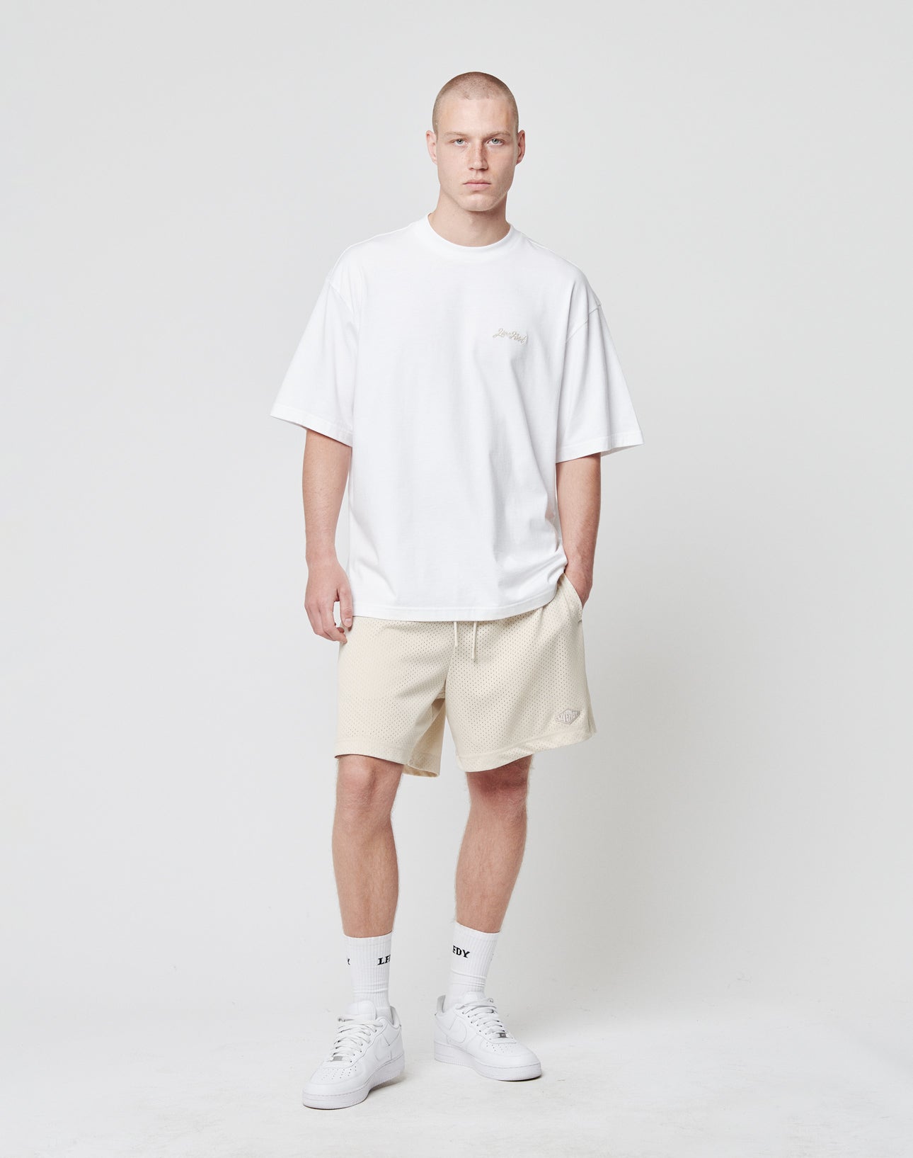 A man with short hair and a neutral expression stands against a white background wearing LIVE FAST DIE YOUNG Basic Mesh Shorts in light beige, a loose Regular Fit white T-shirt, white socks, and white sneakers, with his left hand in his pocket.