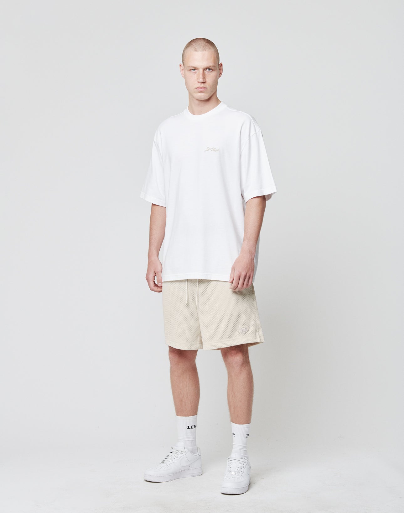 A man with a buzz cut stands in a neutral pose wearing the LIVE FAST DIE YOUNG Basic Script Tee, loose and white, paired with light beige shorts, white socks, and sneakers against a plain light gray background.