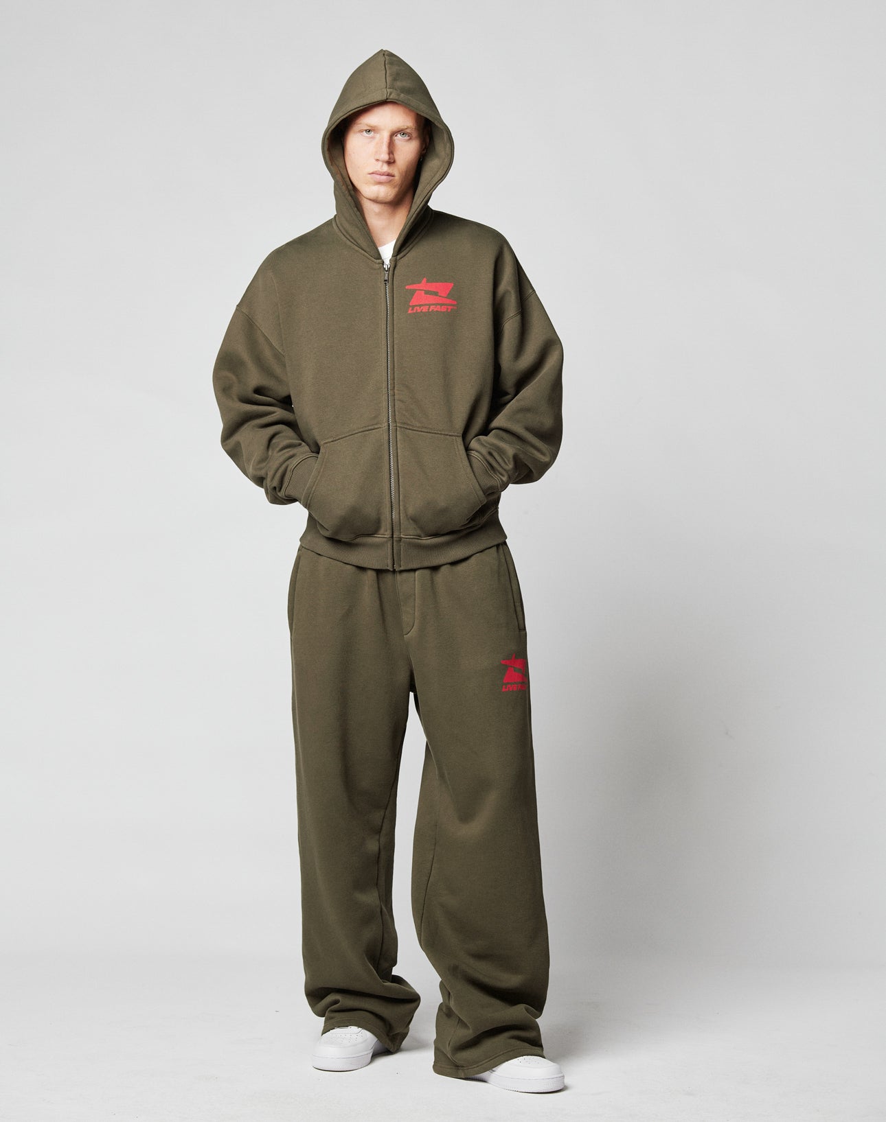 A person wears the LIVE FAST DIE YOUNG Live Fast Sweatsuit Jacket in olive green with a red front logo, hood, pockets, and white sneakers—an oversized fit made from premium cotton.