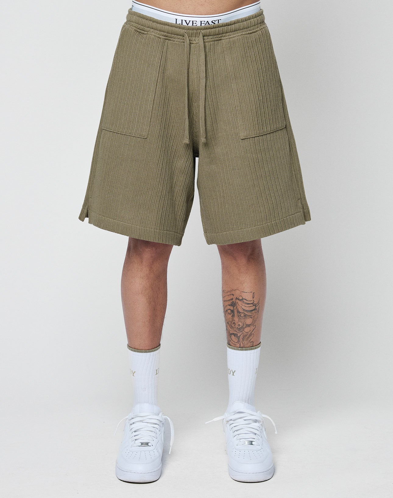 Someone wears olive green Basic Knit Shorts from LIVE FAST DIE YOUNG with a white branded waistband, white socks, and sneakers. Their lower legs, including a left shin tattoo, are visible against a plain background.