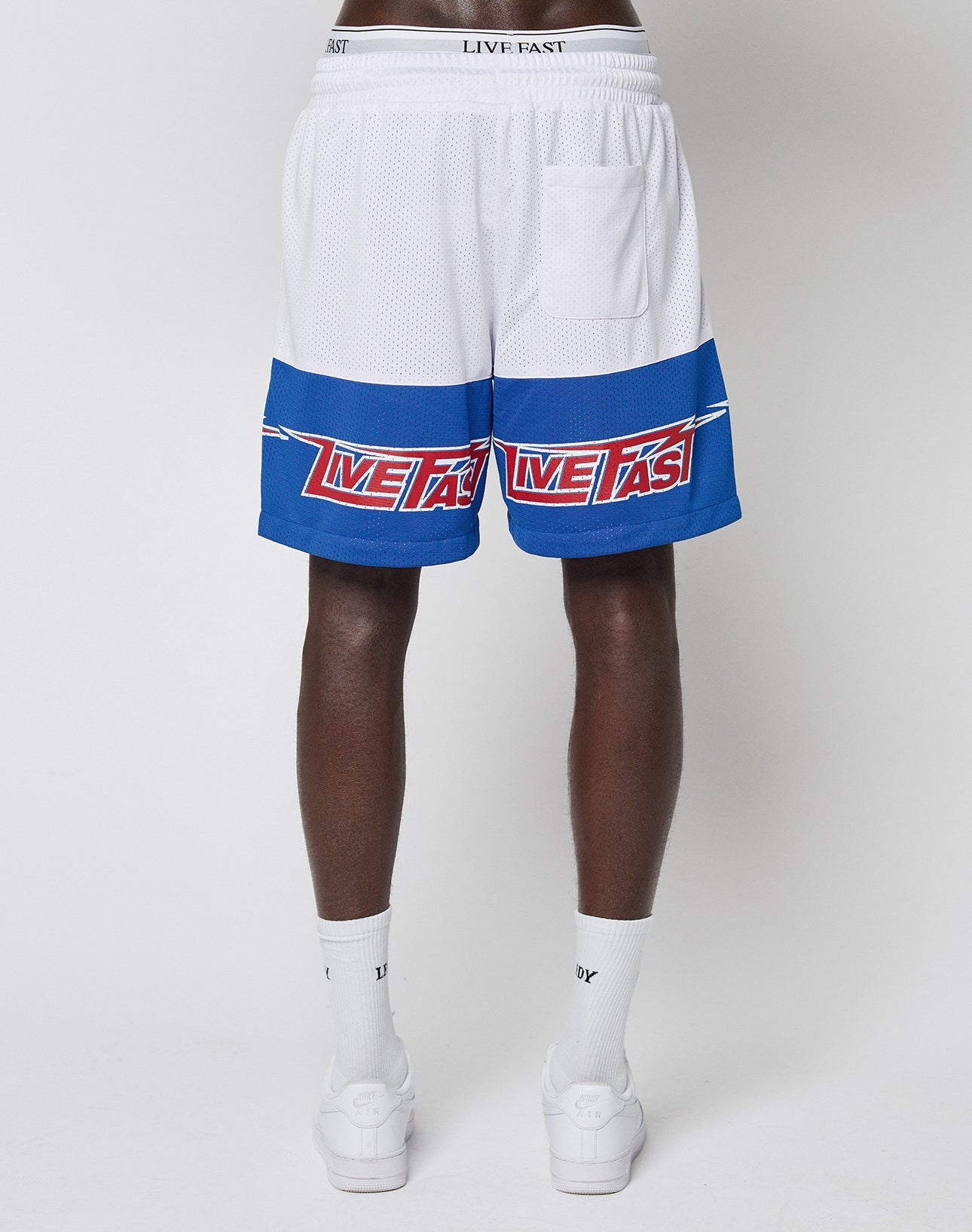 A person stands backward, wearing LIVE FAST DIE YOUNG LFDY Racing Mesh Shorts with blue stripes and red text, featuring großflächige Prints, paired with white socks and slides against a plain white background.