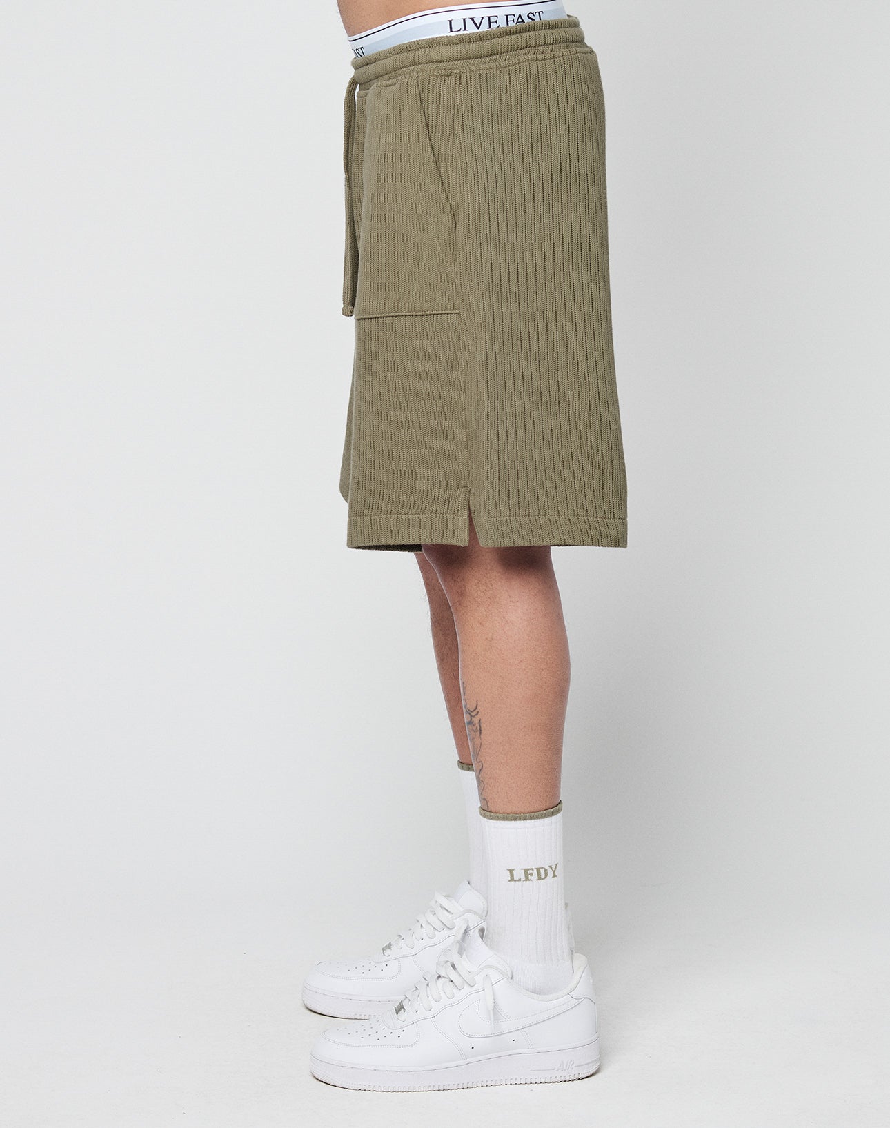 A person in profile wears olive green Basic Knit Shorts by LIVE FAST DIE YOUNG, featuring an elastic waistband with LIVE FAST text, white “LEDY” socks, and white sneakers against a plain white background.