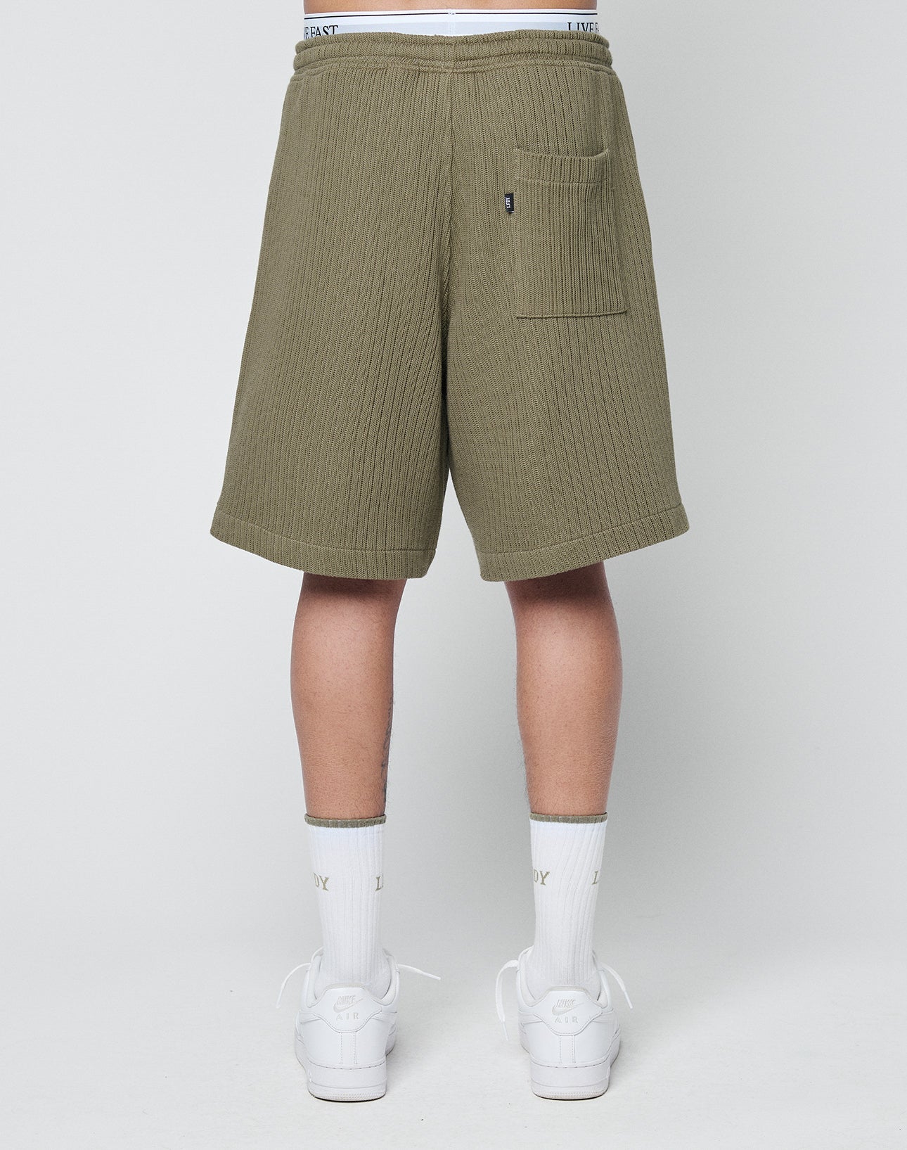 A person stands facing away in olive green Basic Knit Shorts from LIVE FAST DIE YOUNG with ribbed texture, a back pocket, elastic waistband with visible branding, white ENJOY socks, and white sneakers against a plain light background.