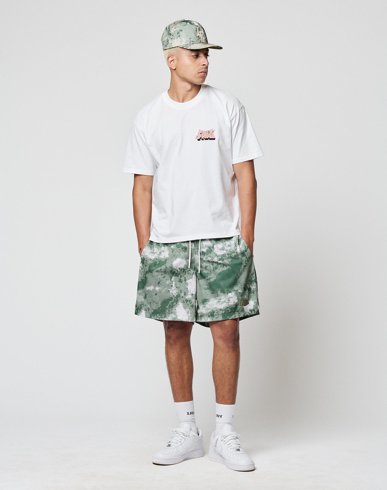 A person stands against a plain light background, wearing a white t-shirt, LFDY Mesh Shorts by LIVE FAST DIE YOUNG in green and white camo, a matching cap, white socks, and white sneakers, looking to the side with hands in shorts pockets.