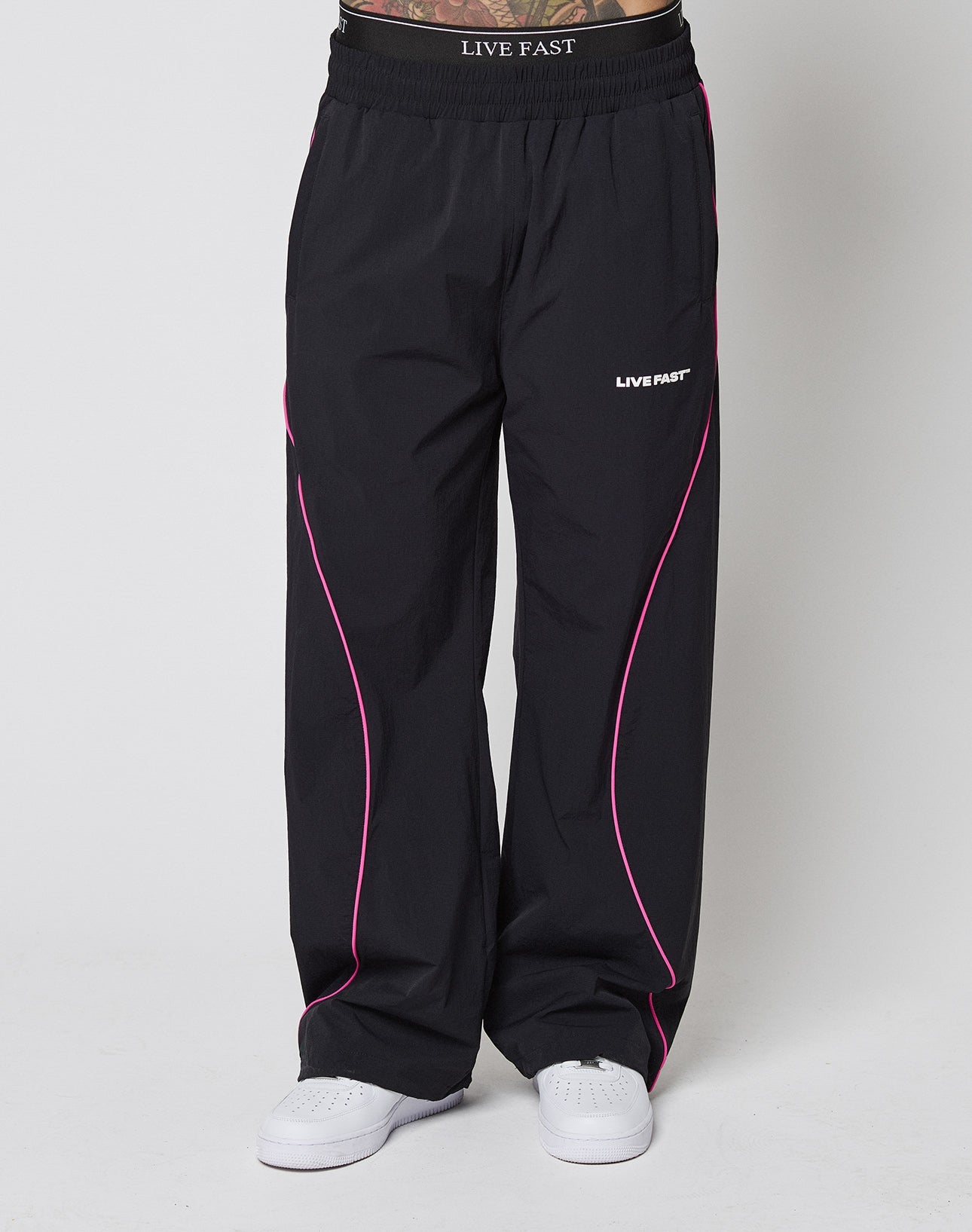 A person wears LFDY Wavy Trackpants by LIVE FAST DIE YOUNG—wide fit, black with pink piping, elastic waistband, and LIVE FAST logos—paired with white sneakers. Only their lower half is visible against a plain background.