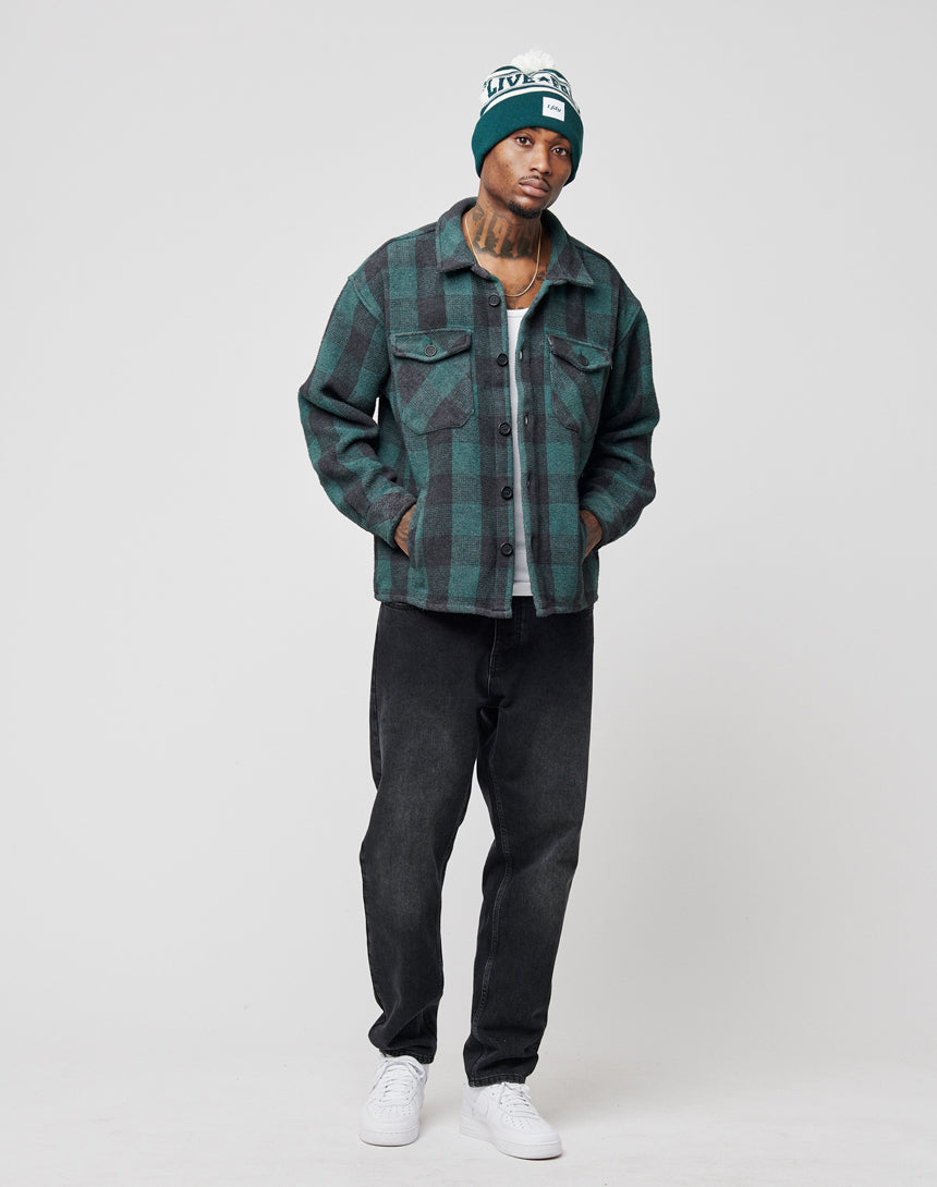 A man wearing a green plaid jacket with lederpatch, white T-shirt, LIVE FAST DIE YOUNG Regular Denim black jeans, white sneakers, and a green beanie stands with hands in pockets against a plain light background.