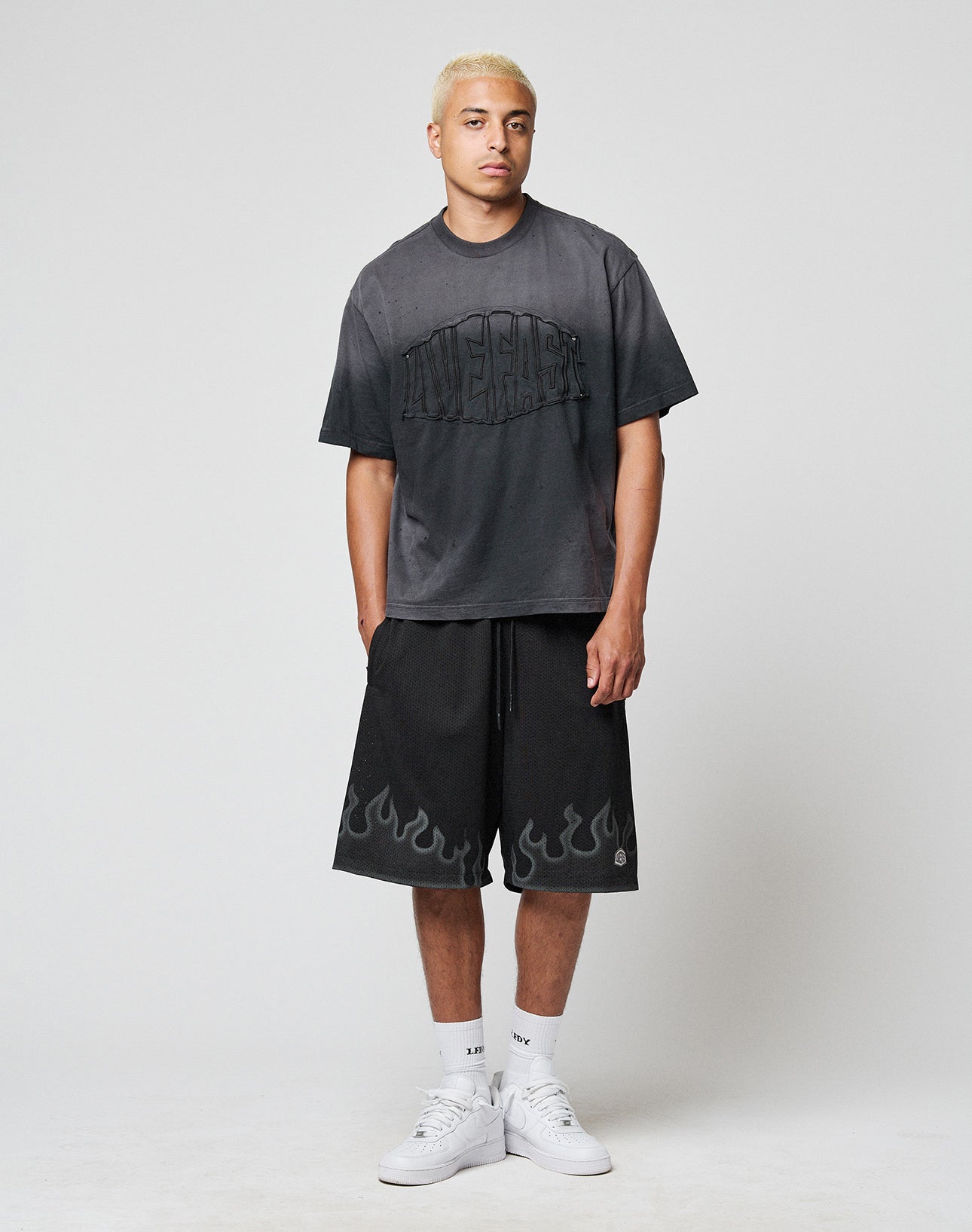 A young man with short blond hair wears a loose black T-shirt, LIVE FAST DIE YOUNG Burning Mesh Shorts with Elastikbund and Flammenprint at the hem, white crew socks, and sneakers, standing against a plain gray background.